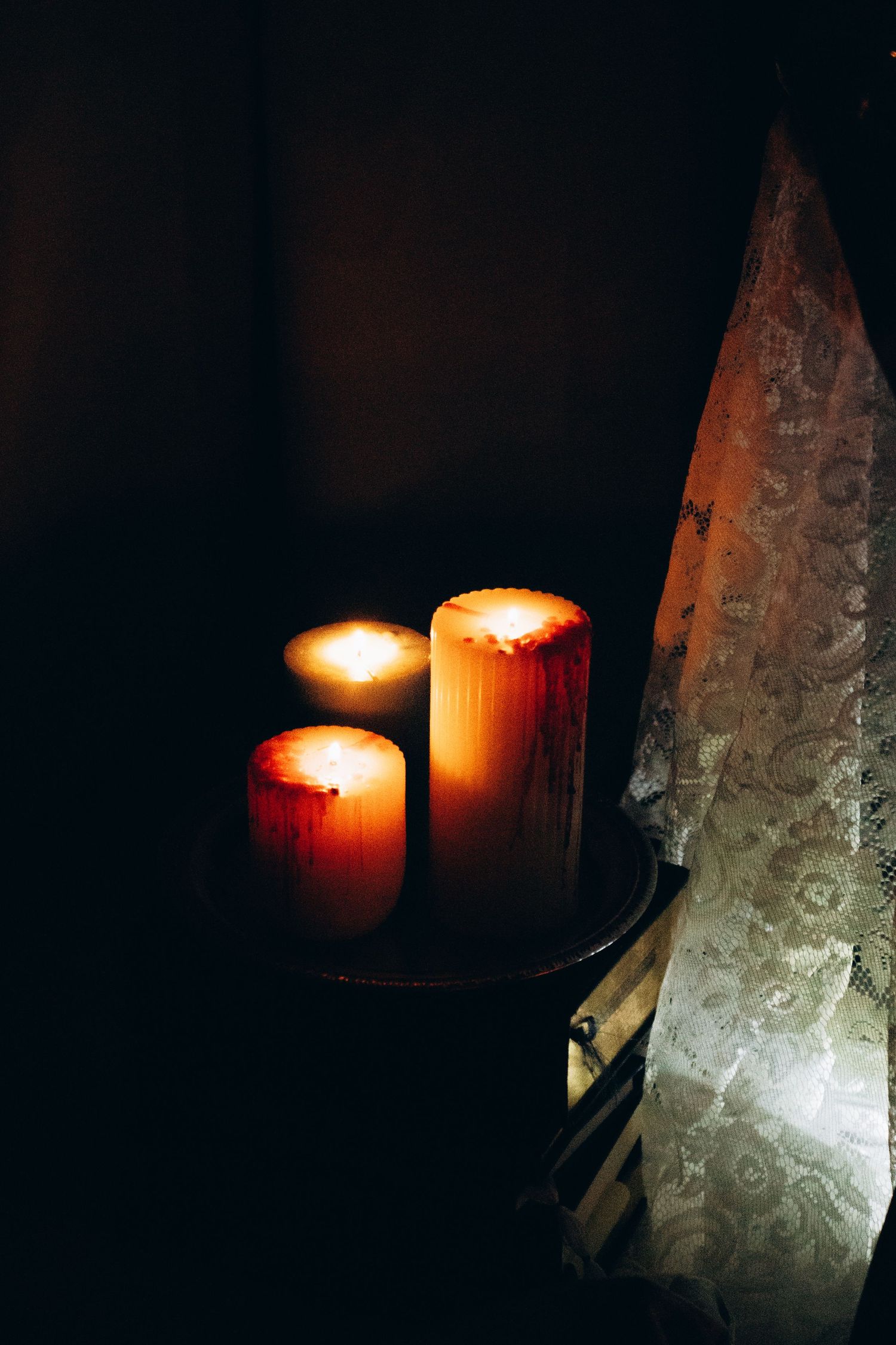 Three glowing pillar candles with red wax drips sit on a dark table beside lace curtains in moody candlelight.