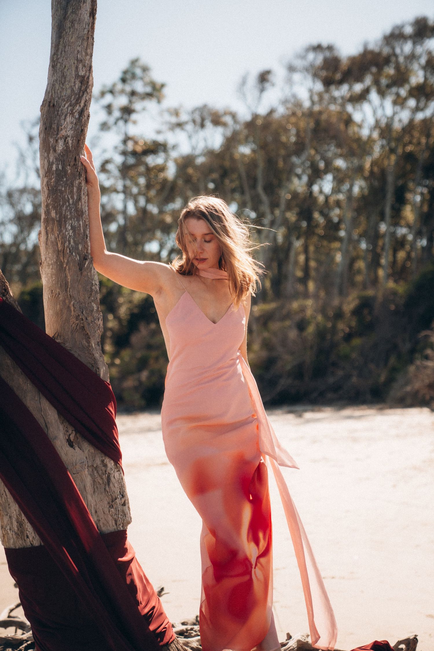 A person in a flowing pink dress dances gracefully on a sandy beach near trees during golden hour.