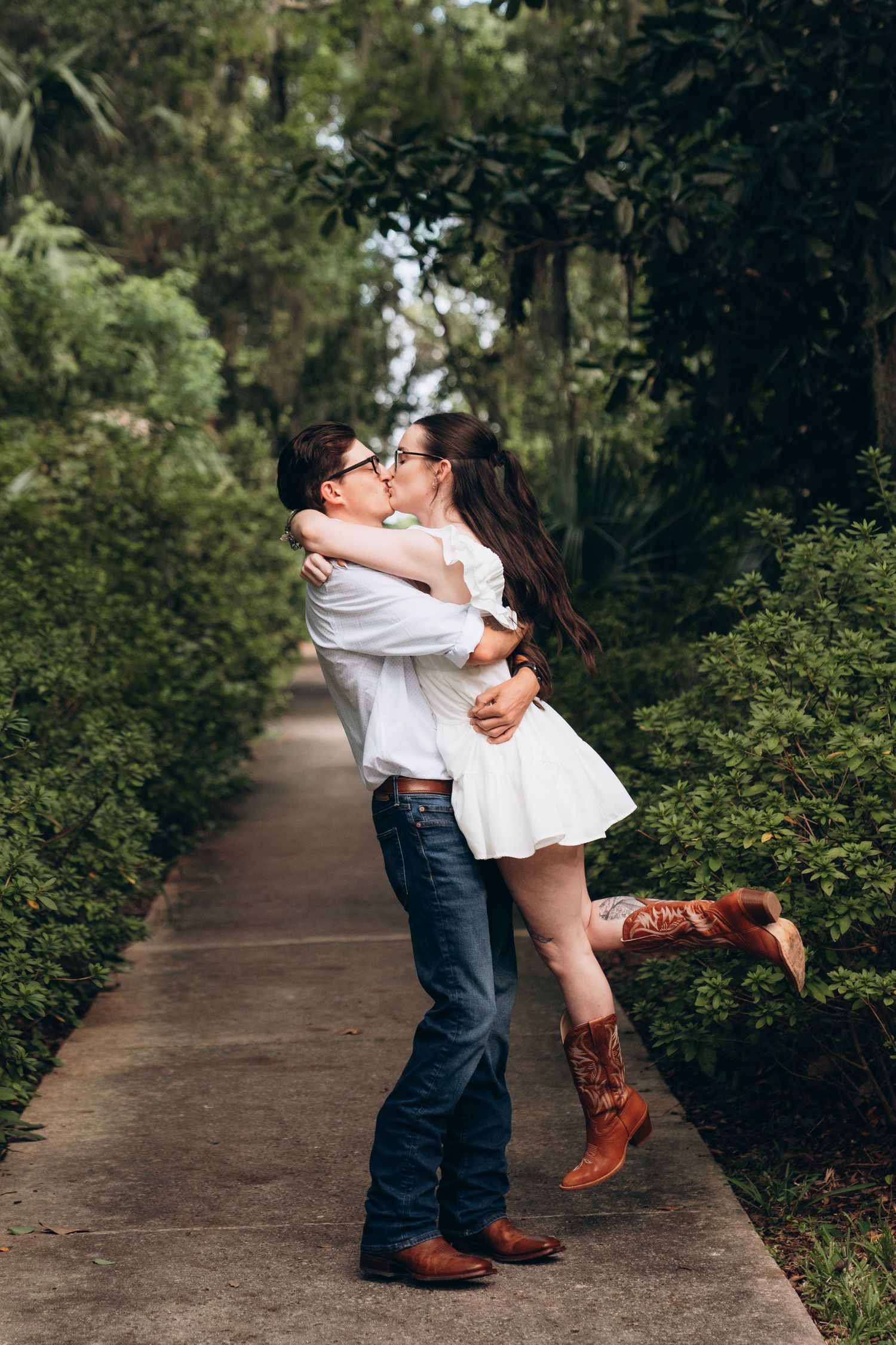 A romantic couple embraces on a garden pathway, with her wearing a white dress and cowboy boots and him in jeans.