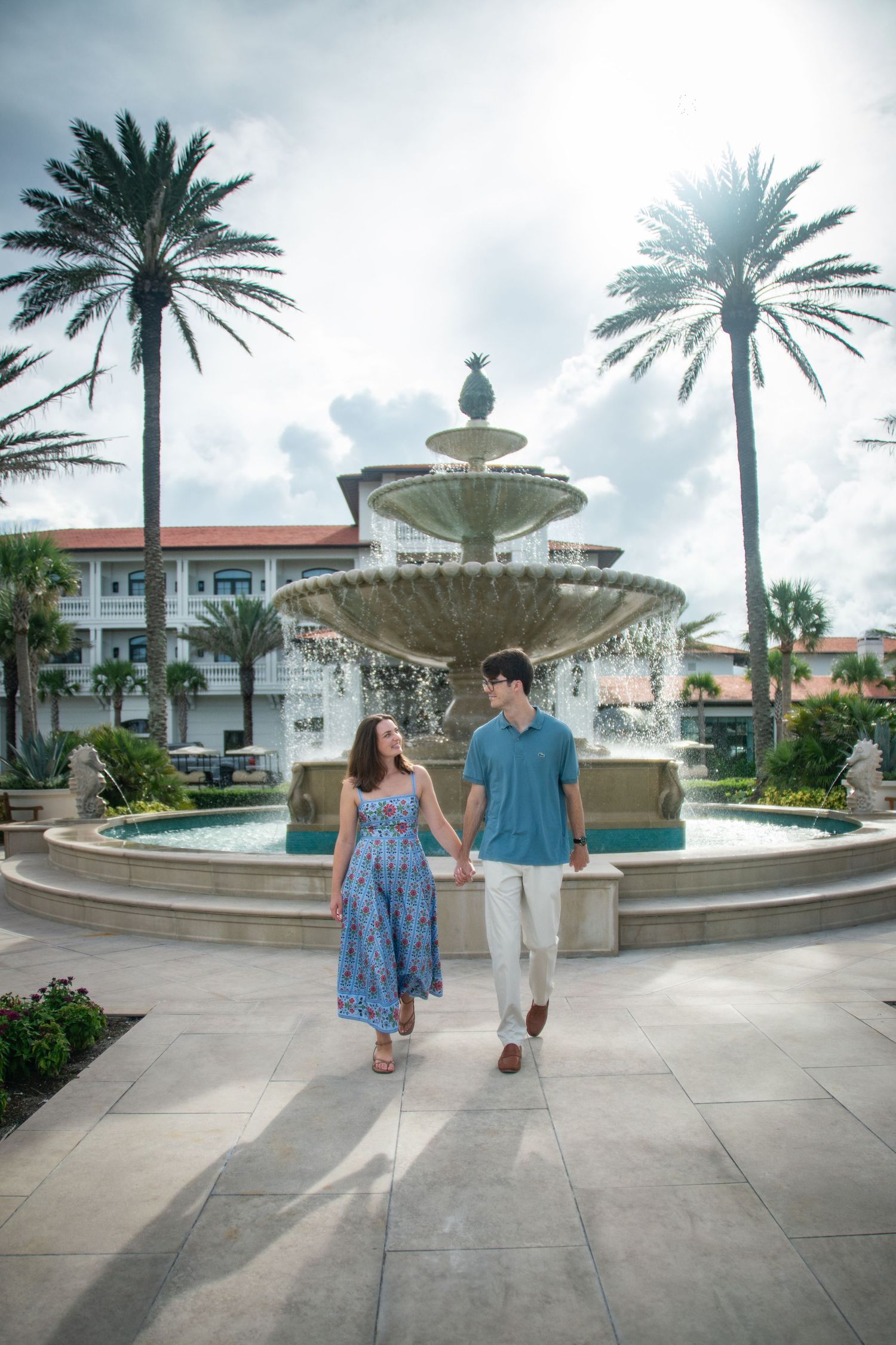 Couple walking past a grand fountain with palm trees and Mediterranean-style architecture at a luxury resort plaza.