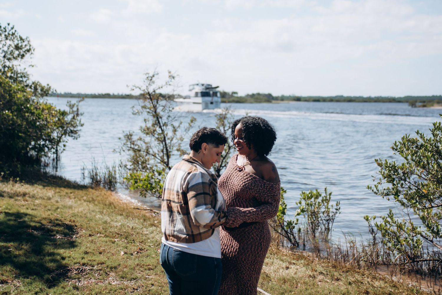 Two people embrace while standing on a scenic waterfront overlook with boats in the distance.