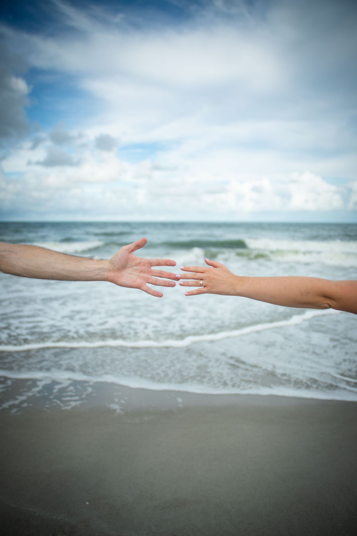 Two hands reach towards each other against an ocean backdrop with waves rolling onto the shore.