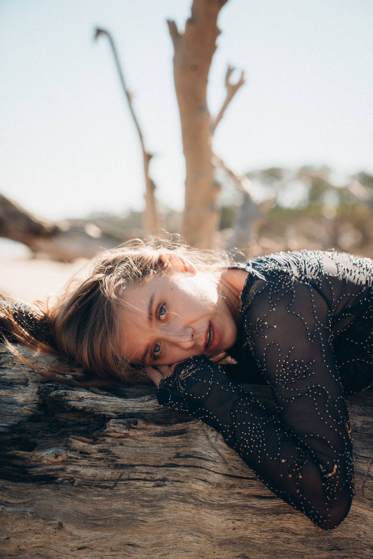 Artistic portrait of someone laying on driftwood at sunset wearing a black sparkly outfit.