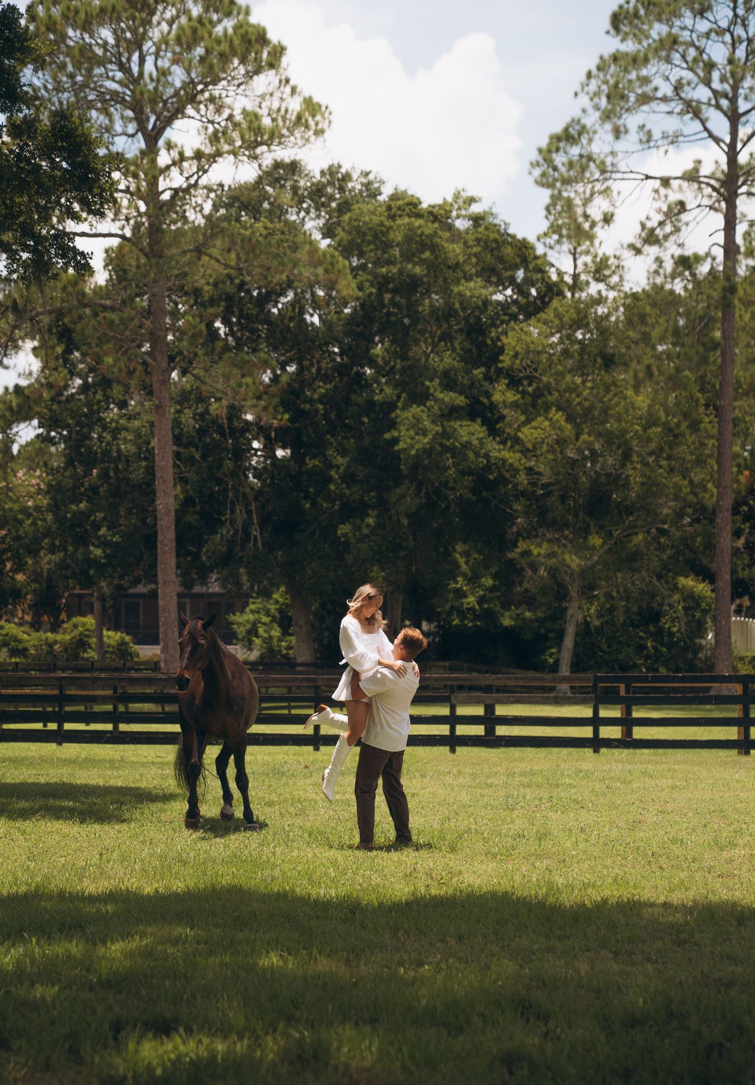 A couple shares a tender moment in a grassy field with a horse grazing nearby.