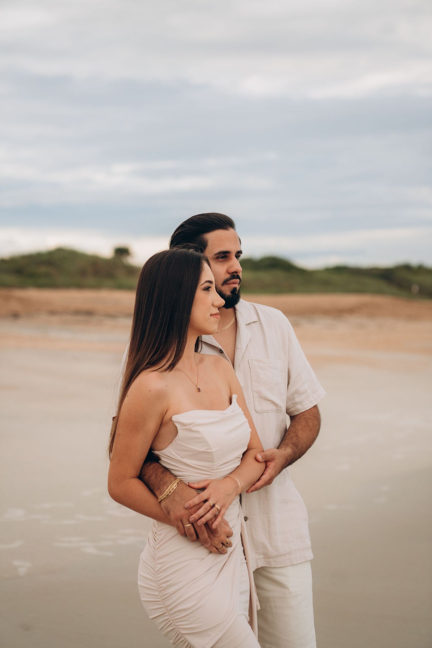 Couple in light colored outfits embracing on sandy beach during overcast day.