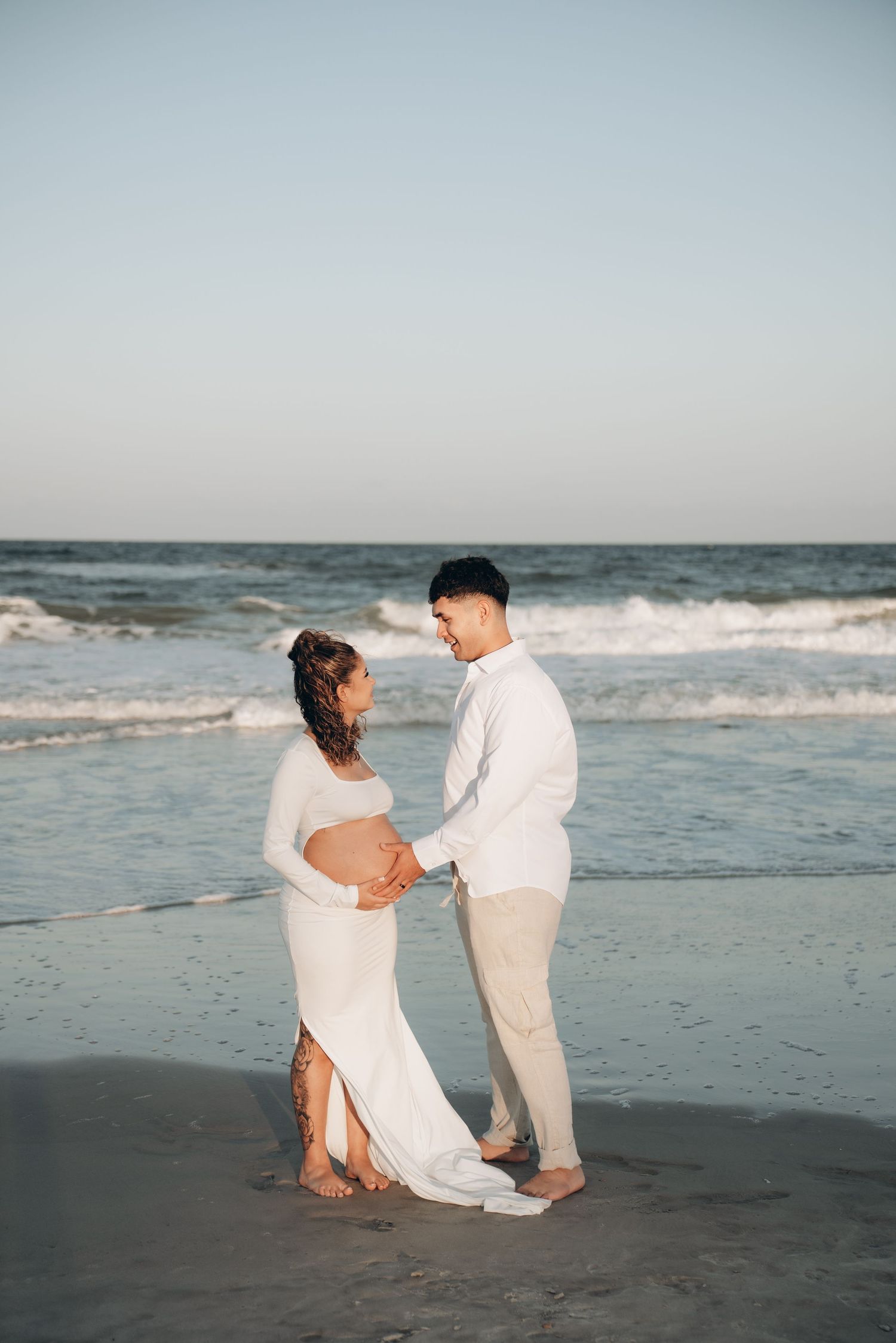 Couple in white attire poses for romantic maternity photos along the shoreline.