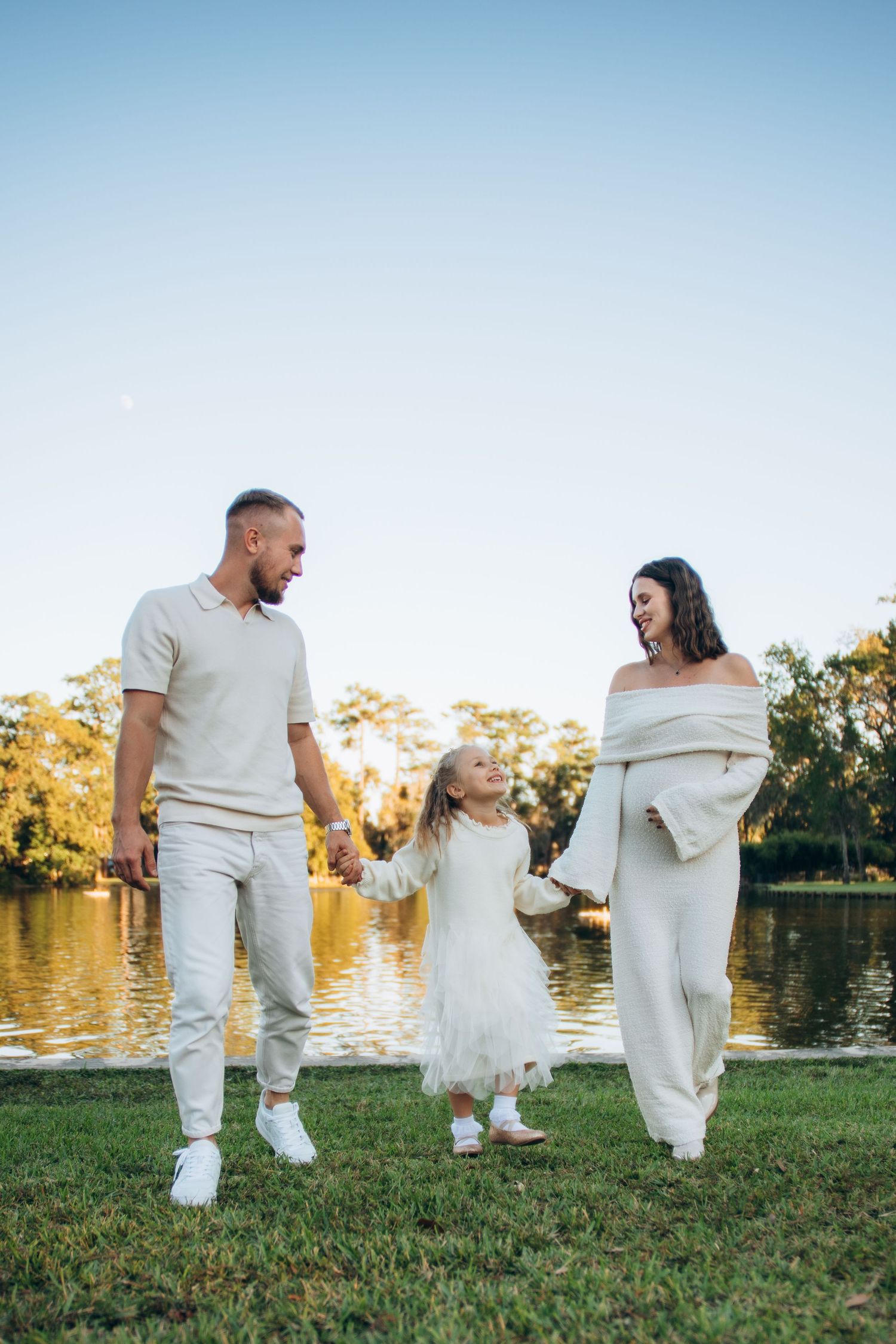 A family in coordinated white outfits walks hand in hand along a lakeside park at sunset.