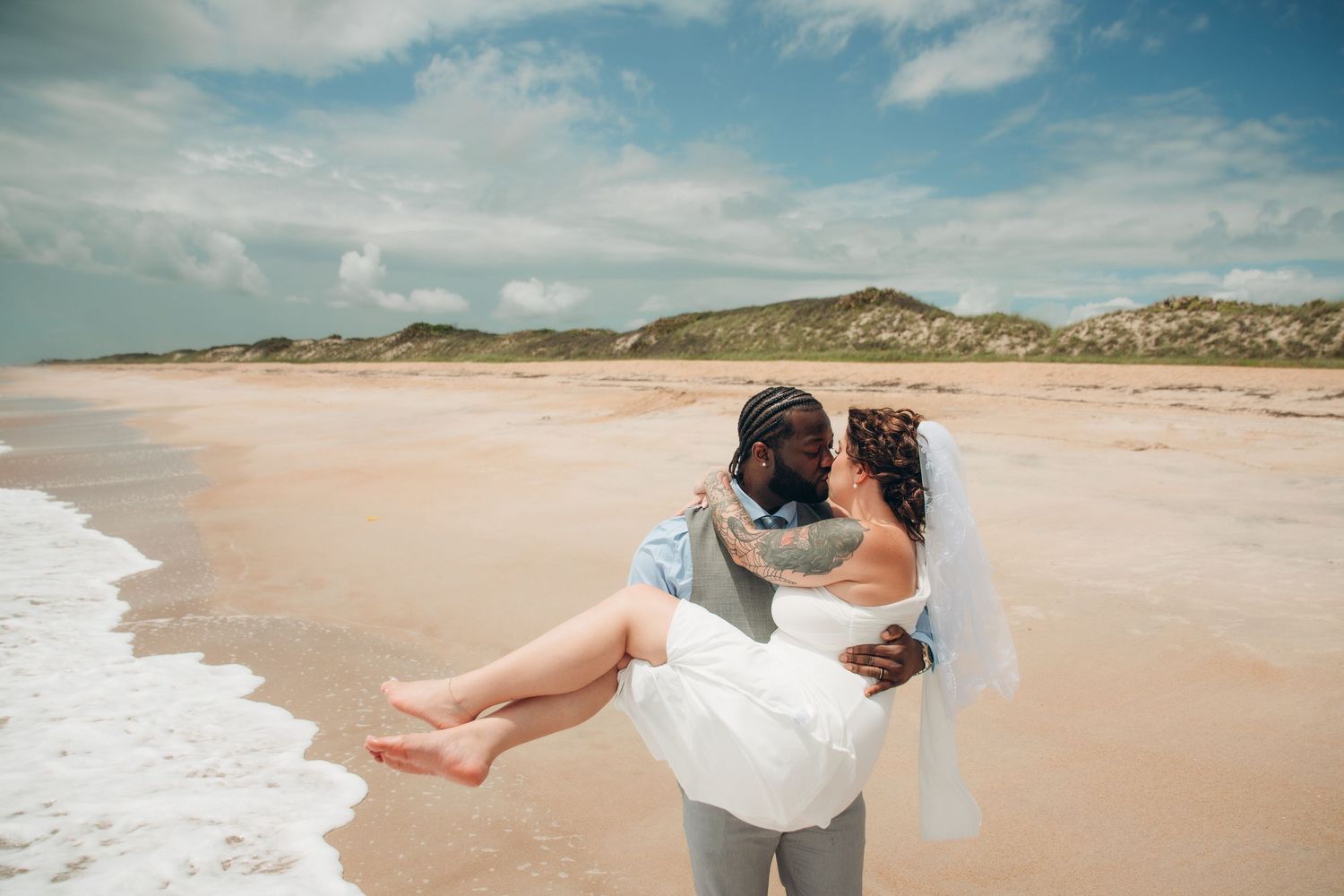 Groom carries bride in white dress and veil on a sunny beach with ocean waves and sand dunes.