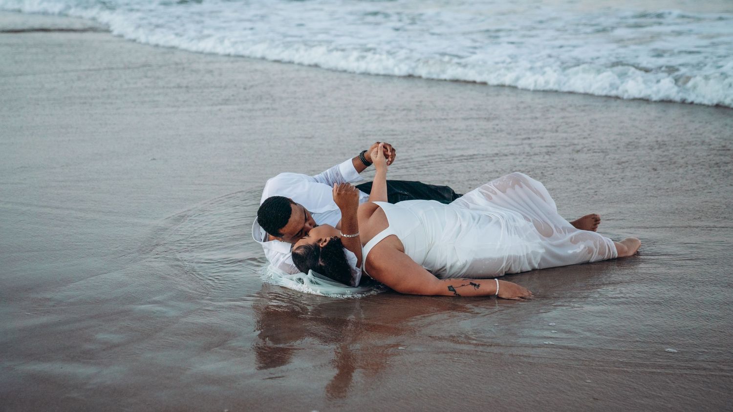 Romantic couple kissing while lying on a sandy beach with ocean waves, bride in white dress and groom in casual attire.