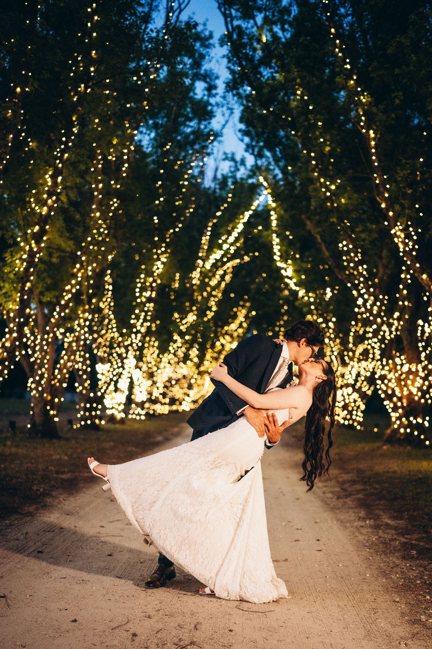 Bride and groom share a romantic dip kiss surrounded by twinkling fairy lights on trees at twilight wedding.