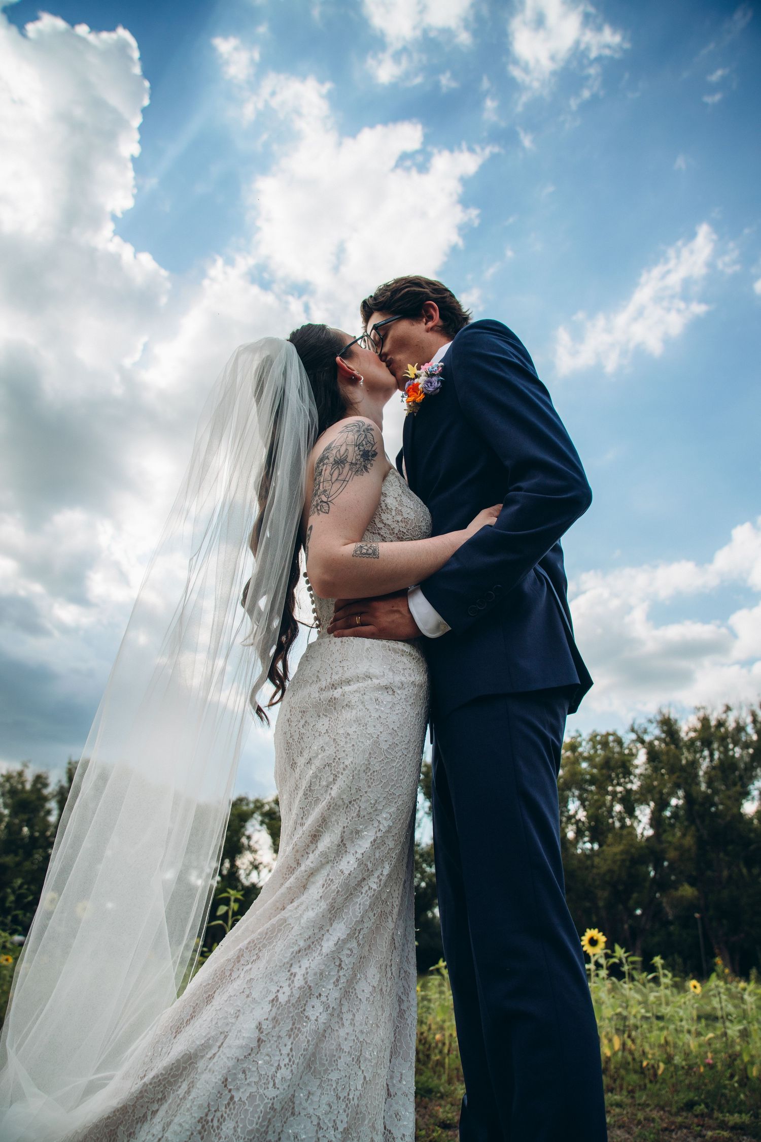 A bride and groom share a romantic kiss outdoors under a blue sky, she wearing a lace gown and veil, he in a navy suit.