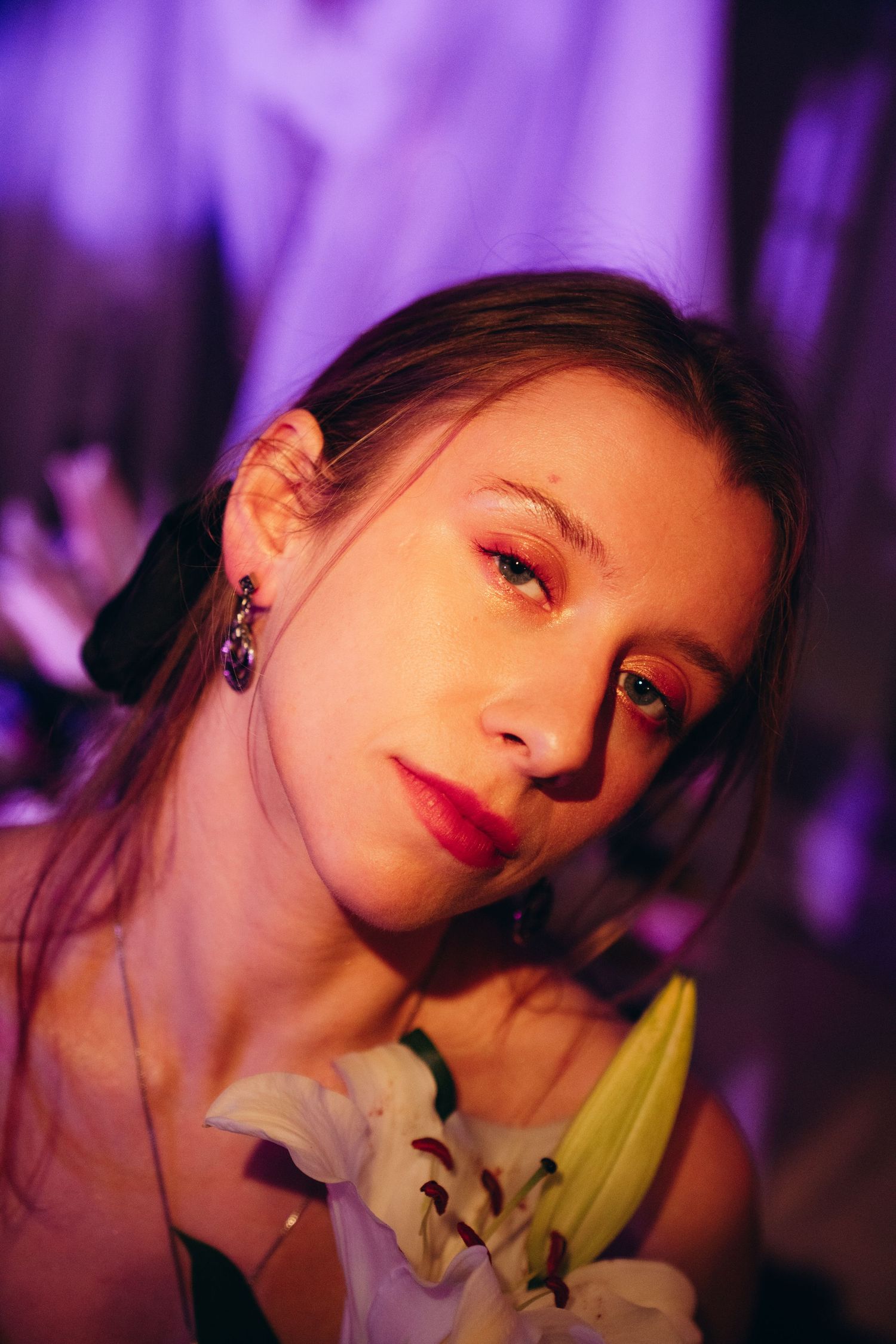 Close-up portrait of young woman with red lips holding white lily under purple ambient lighting.