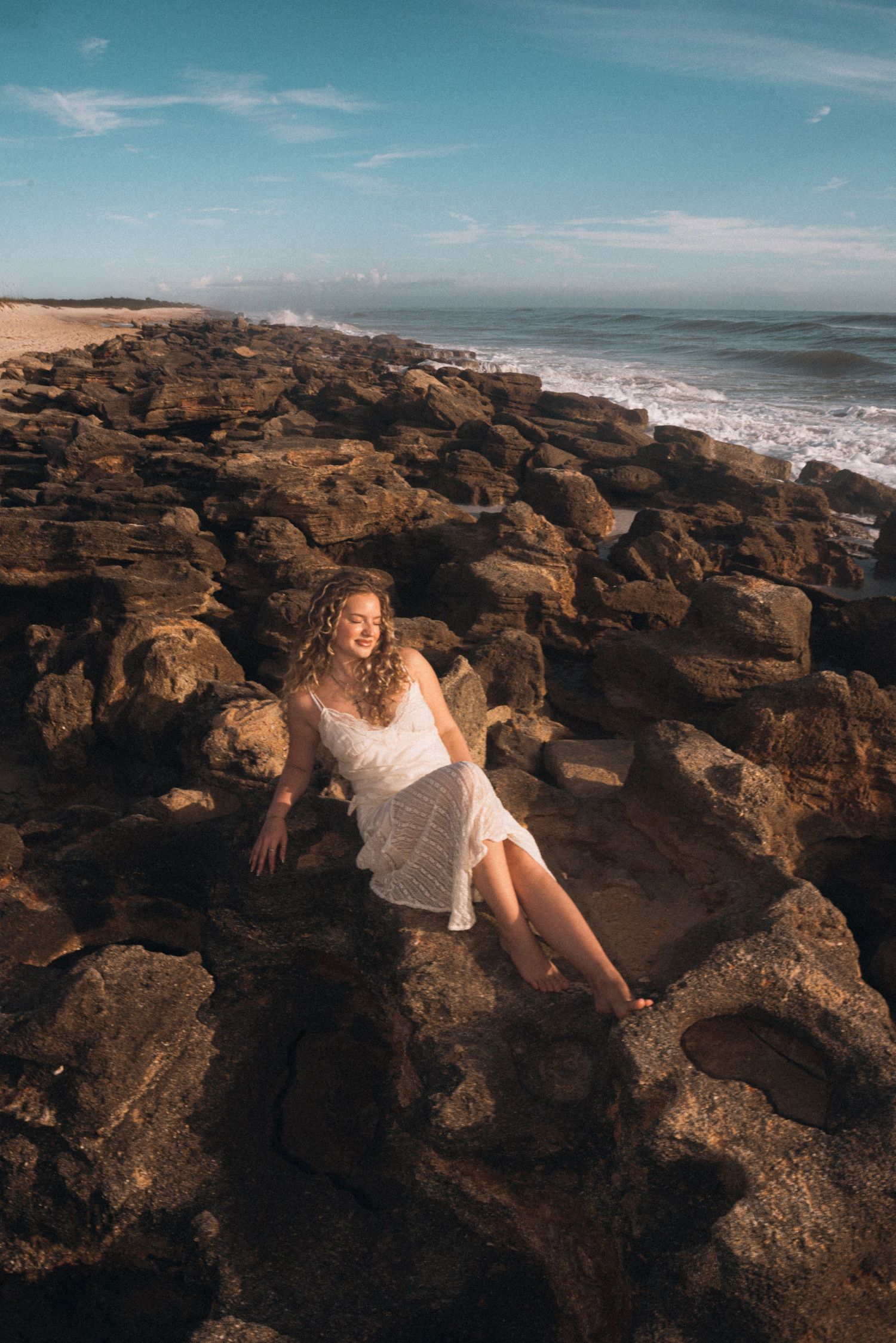 Woman in white lace dress sits gracefully on rocky coastal shoreline with ocean waves in background.