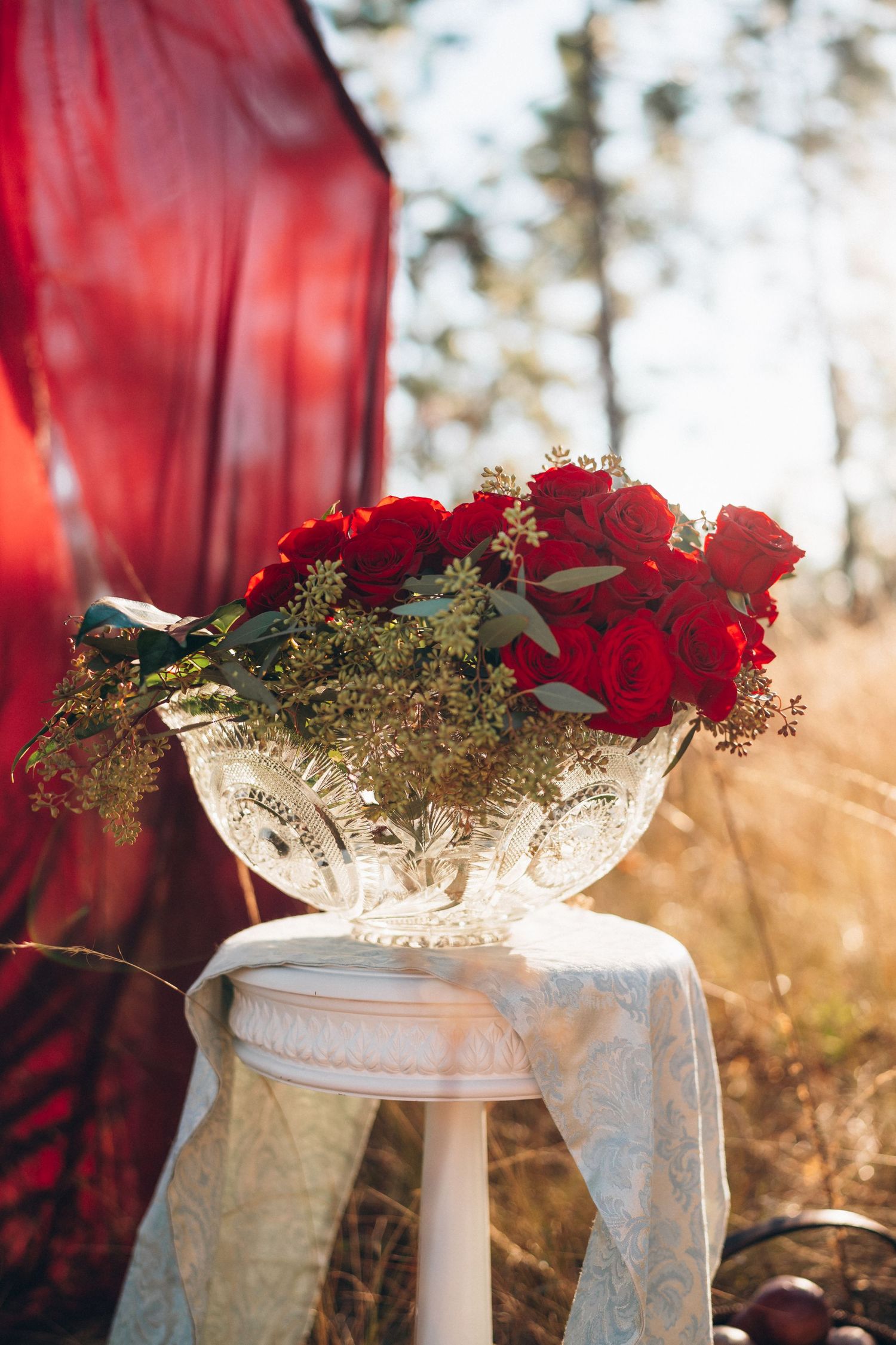 Red roses in crystal bowl on white stool outdoors with red drape backdrop.
