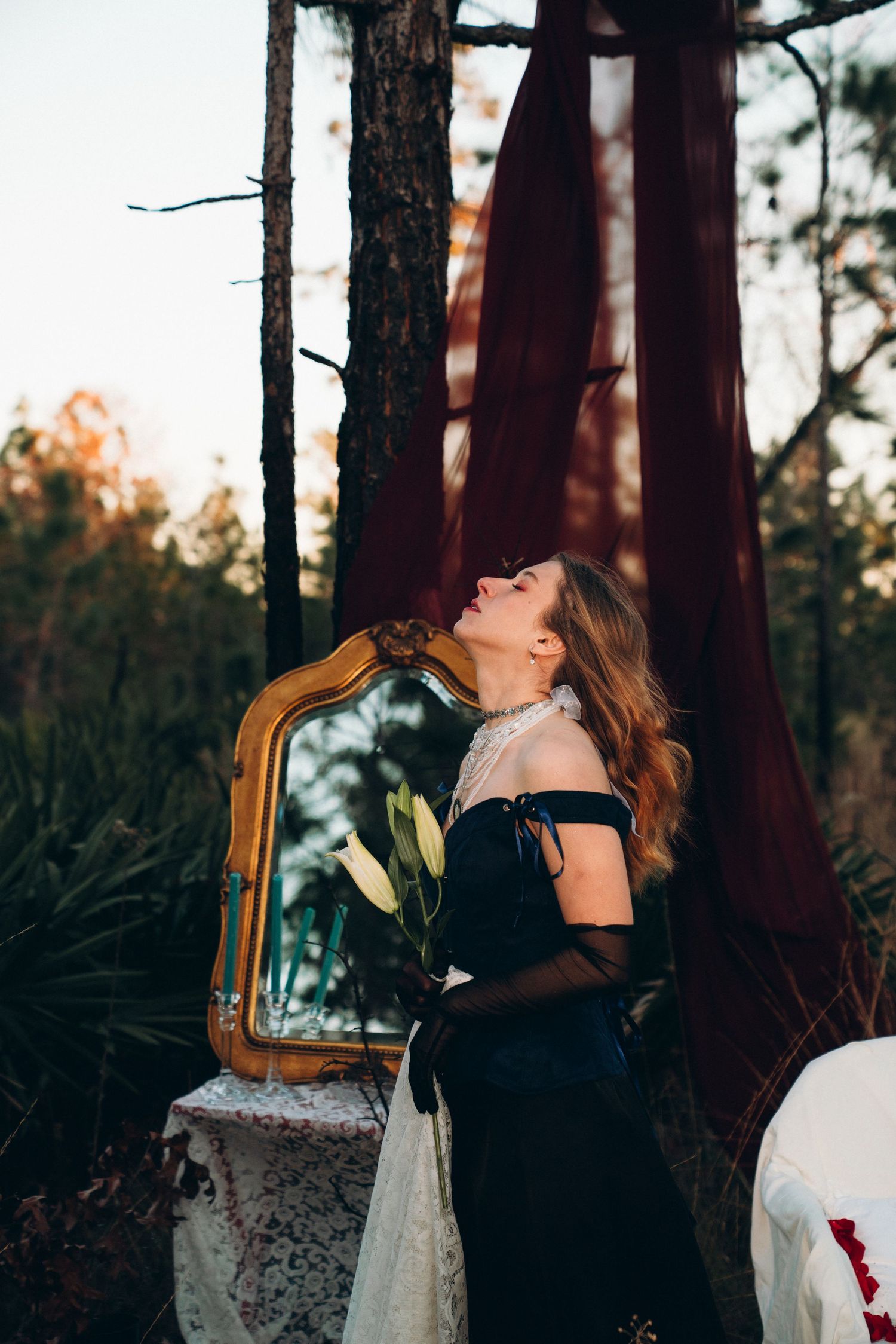 Woman in black off-shoulder gown holding white flowers beside ornate mirror in forest setting.