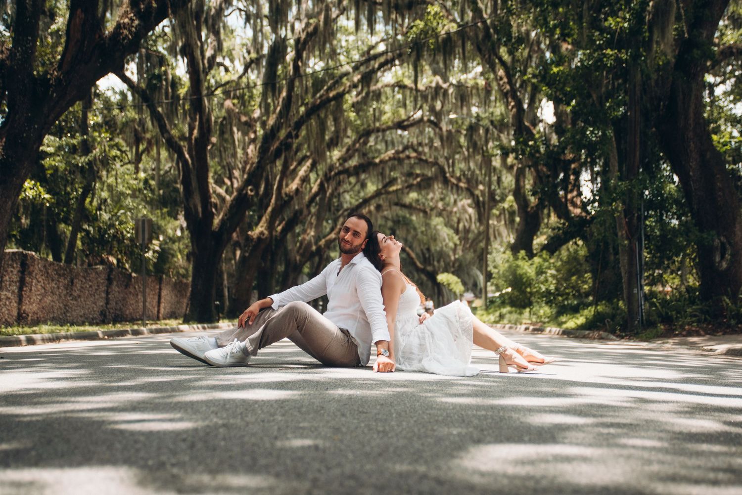 Couple sitting back-to-back on a tree-lined road, dressed in white outfits, surrounded by lush green trees.