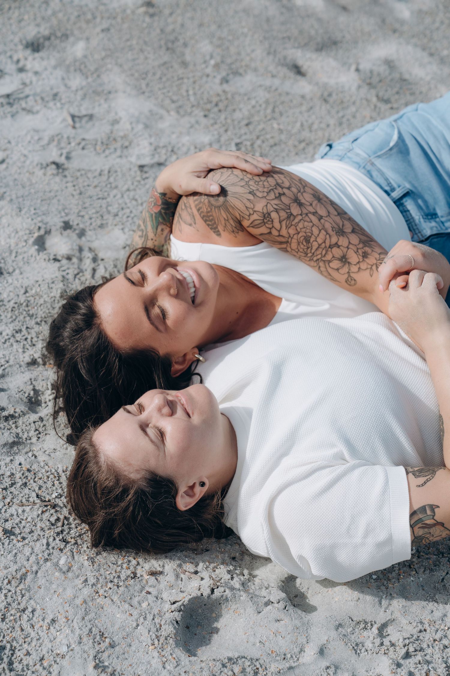 Two people lying on sandy beach wearing white and denim casual clothing.