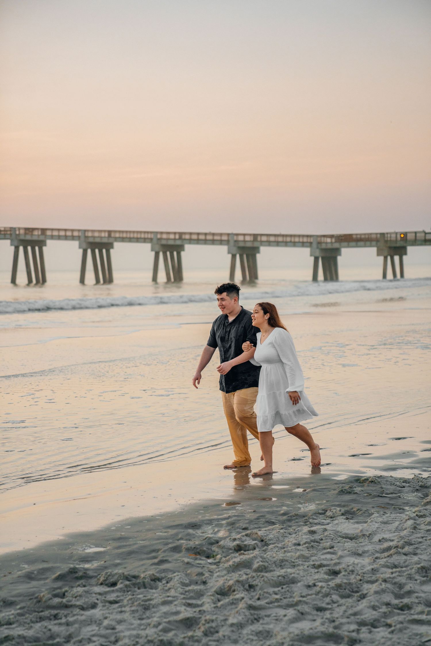 A couple walks along the shoreline with a pier visible in the background at dusk.