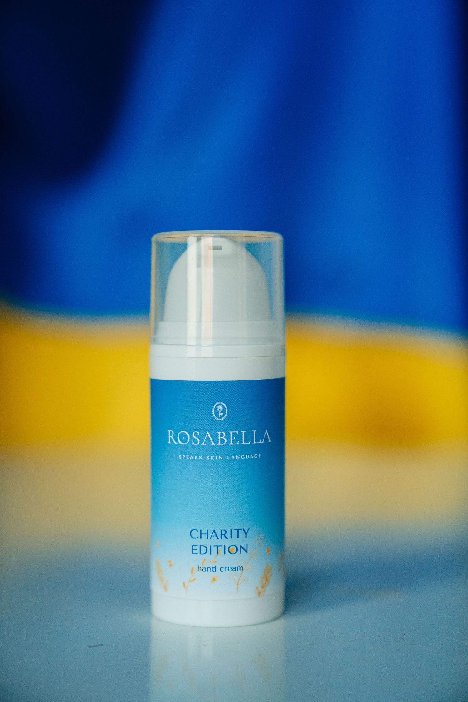 Rosabella Charity Edition hand cream in a pump bottle against a blue and yellow Ukrainian flag background.