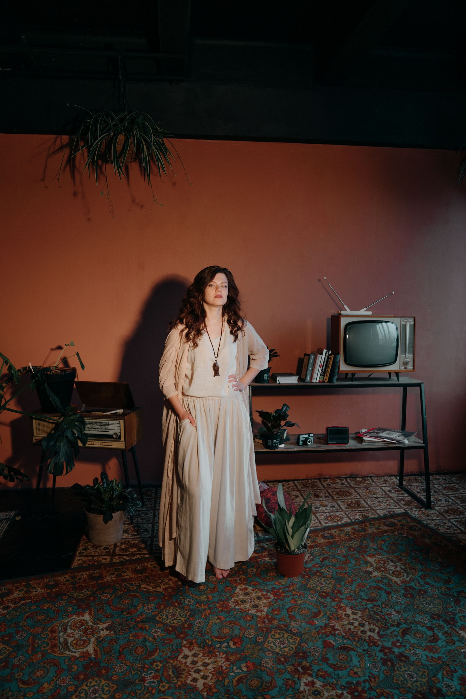 Woman in cream maxi dress poses in vintage-styled room with retro TV, patterned rug, and terracotta walls.