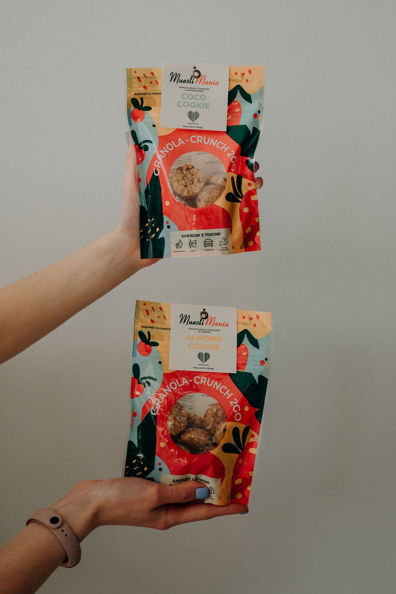 Two hands holding colorful MuesliMania Granola Crunchco snack packages with festive fruit-themed packaging design.