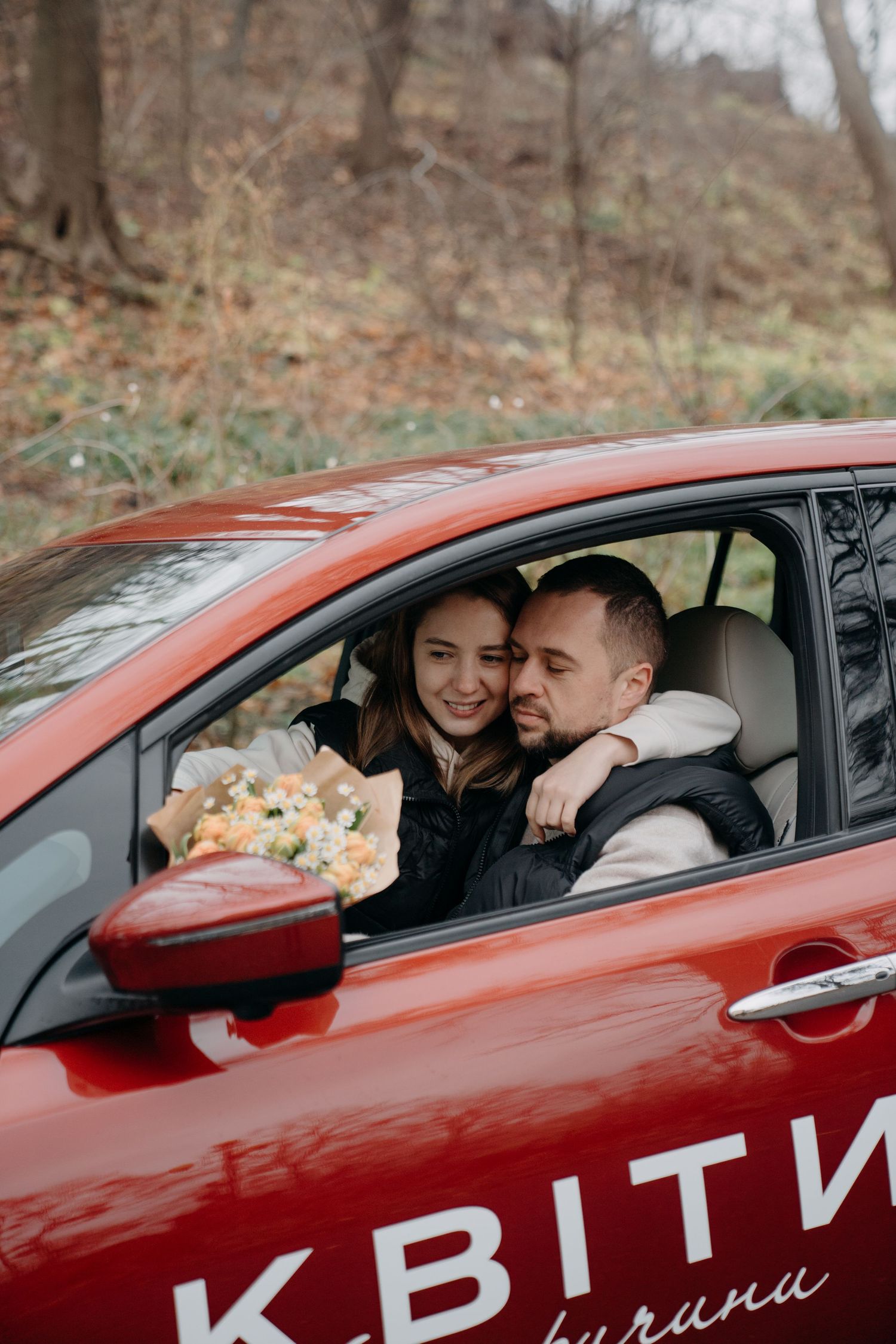 Smiling couple embracing inside a red car with a bouquet of flowers on the window.