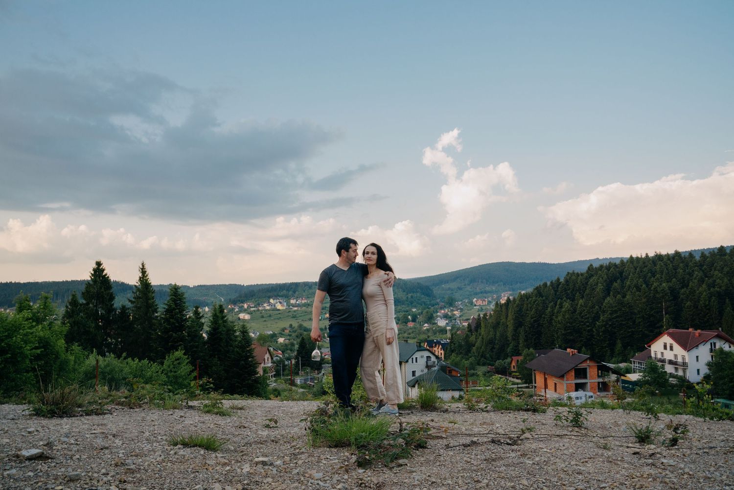 Couple embracing on a hilltop with lush green forest, village houses, and dramatic cloudy sky in background.