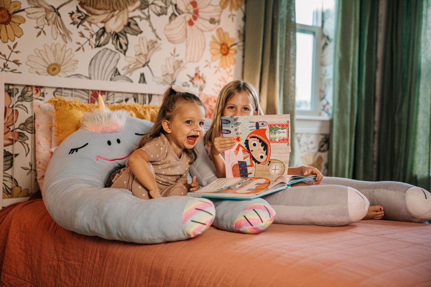 Two young girls sitting on a bed reading a book with a unicorn plush pillow in a floral bedroom.