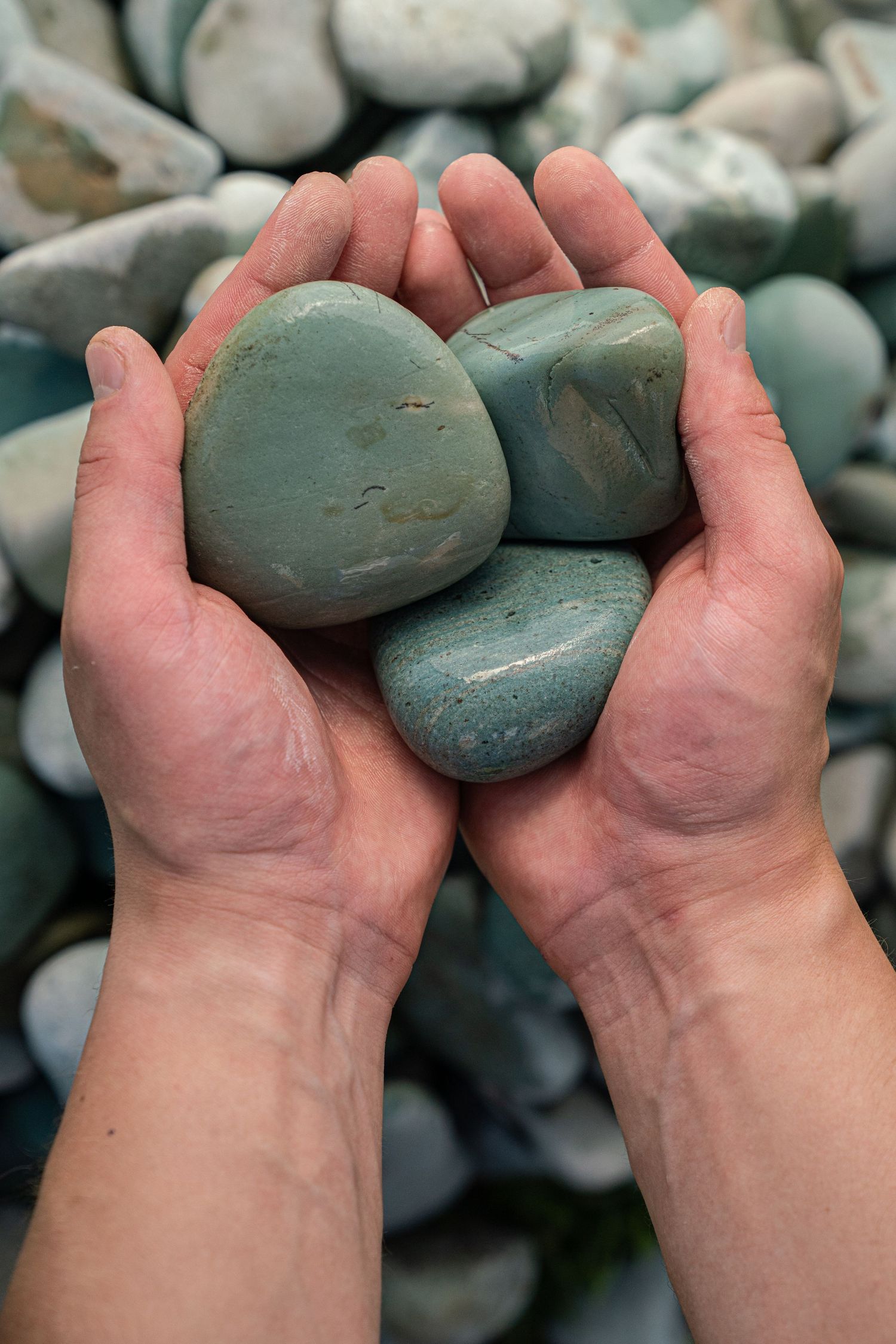 Hands holding three smooth teal green polished stones against a background of similar colored pebbles.