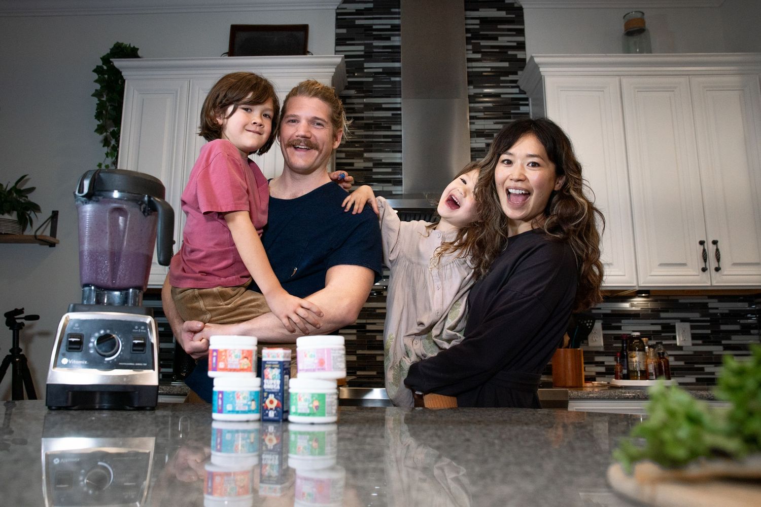 Happy family of four laughing together in modern kitchen with health supplements on granite countertop.