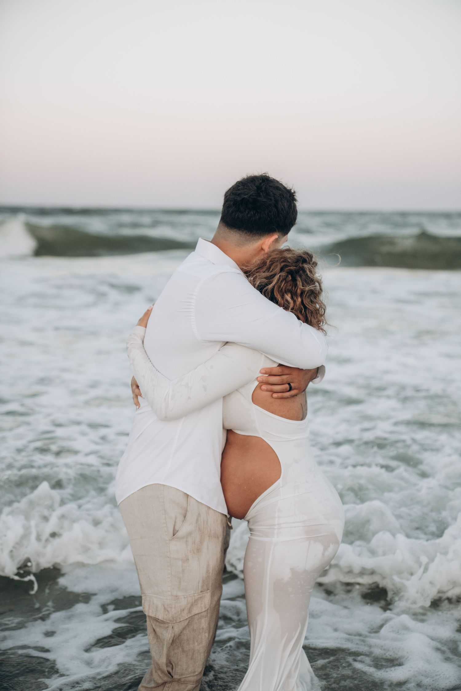 A couple embraces while ocean waves crash around them during a beach photoshoot.