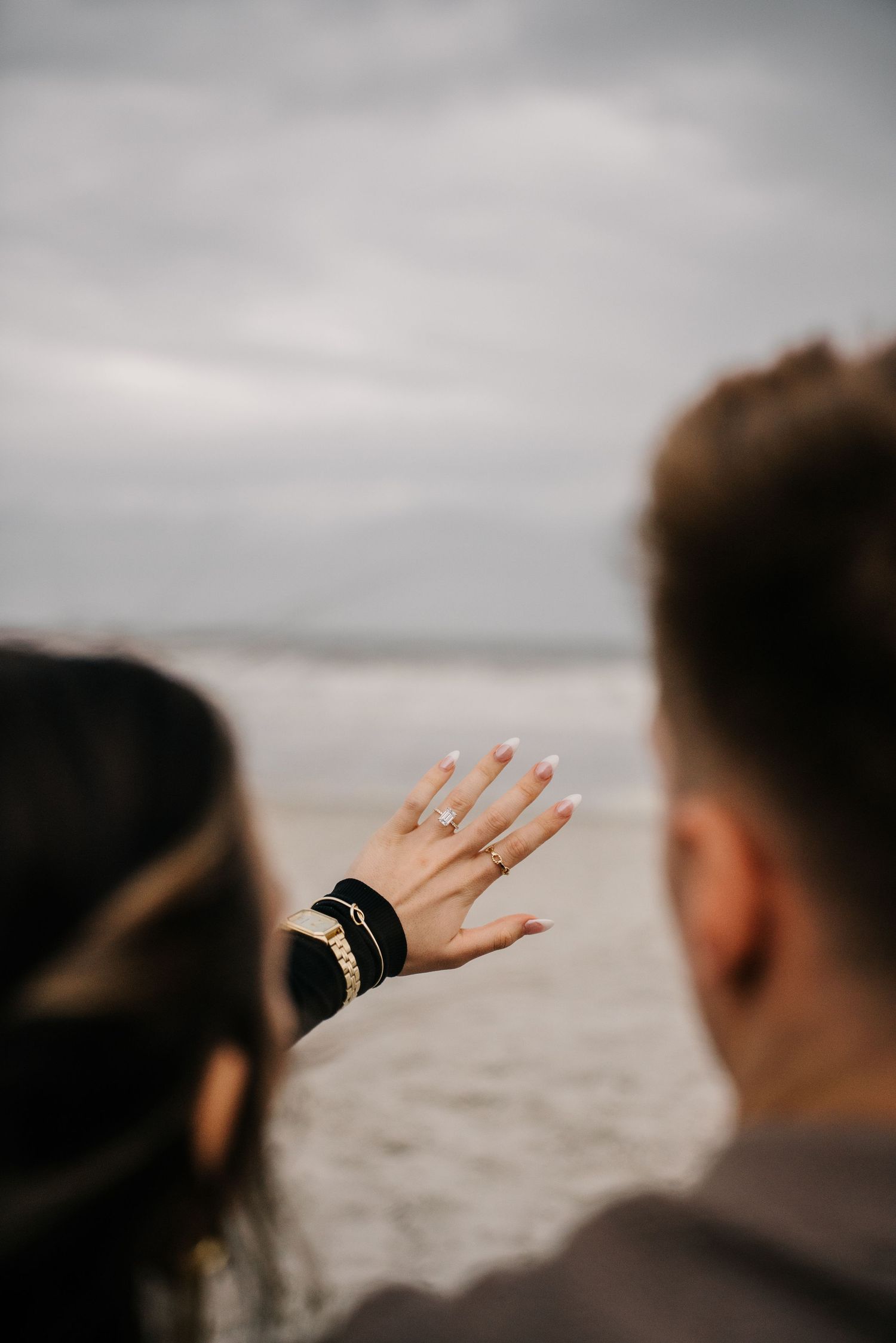 A close-up shot of hands displaying an engagement ring against a cloudy beach background.