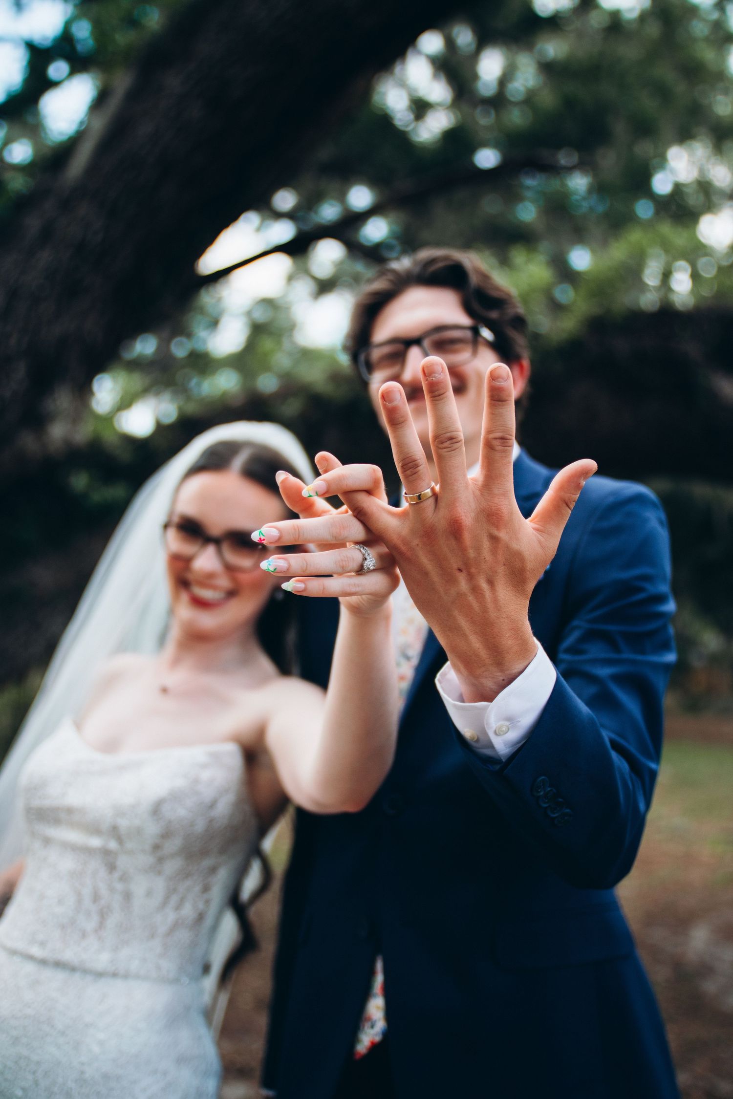 Bride and groom showing off wedding rings outdoors, both wearing glasses, bride in white gown with veil, groom in blue suit.