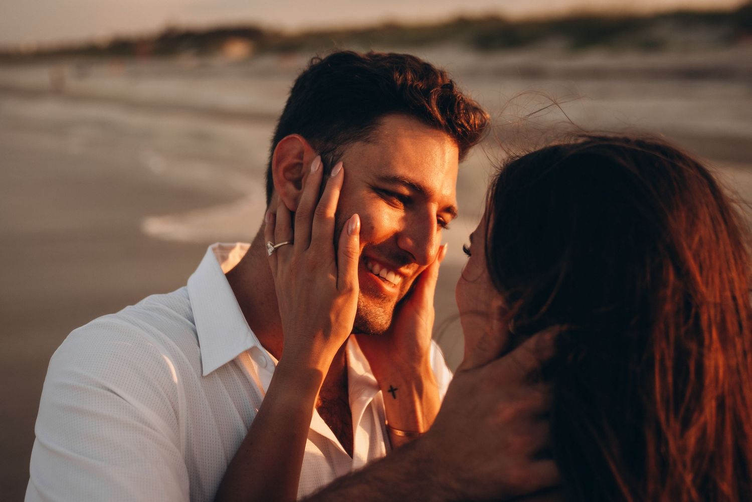Couple sharing a romantic moment on the beach at sunset, woman touching mans smiling face with engagement ring visible.