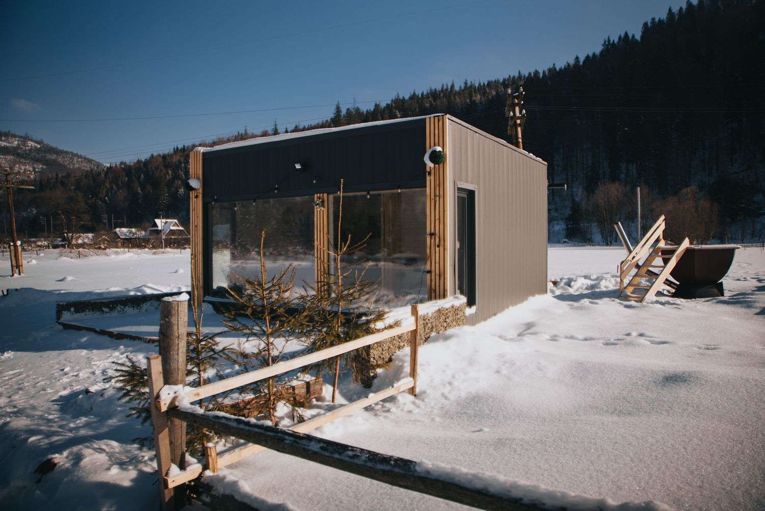 Modern modular cabin in snowy mountain landscape with pine forest backdrop on a clear winter day.