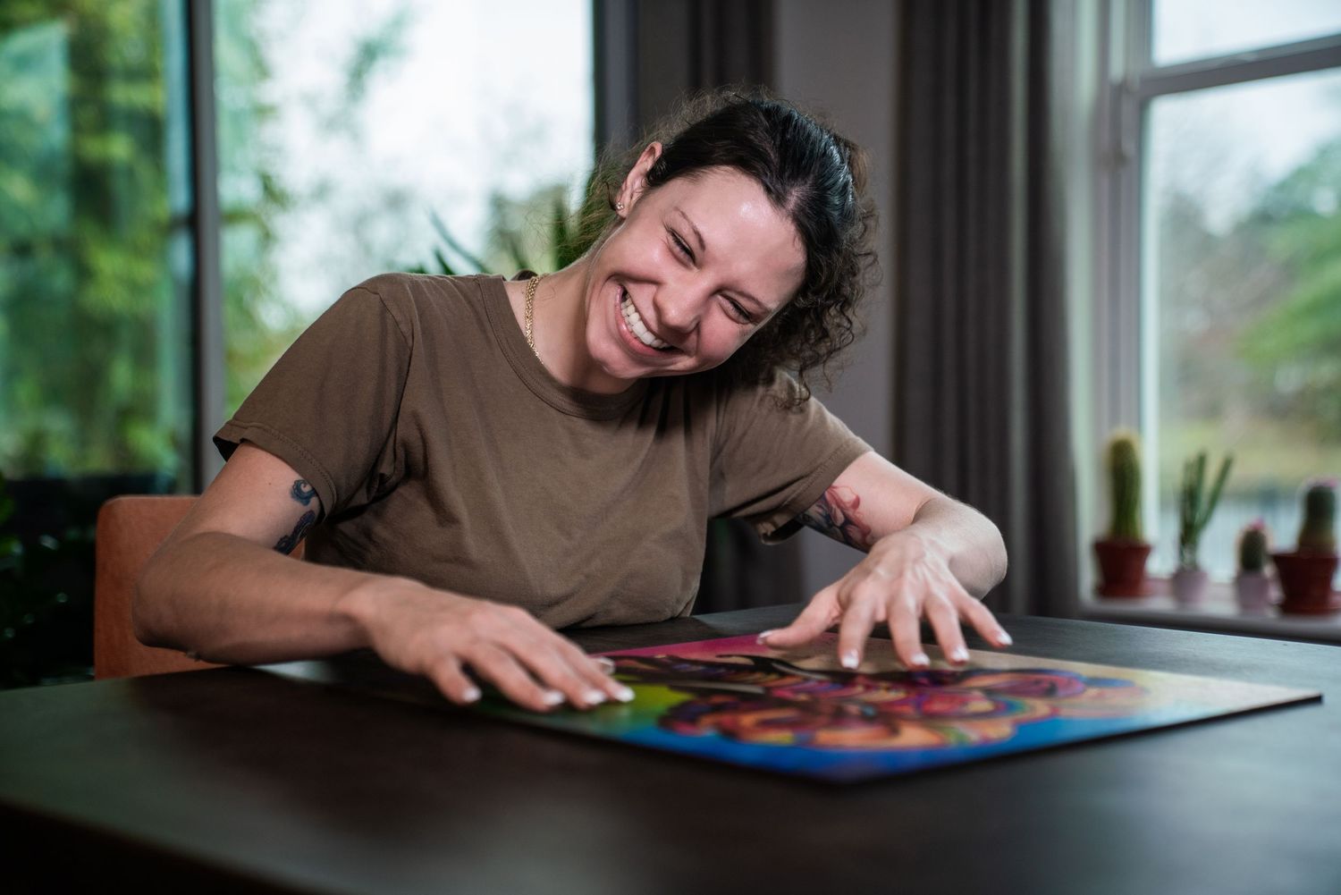 Person smiling while assembling a colorful jigsaw puzzle at a wooden table indoors.