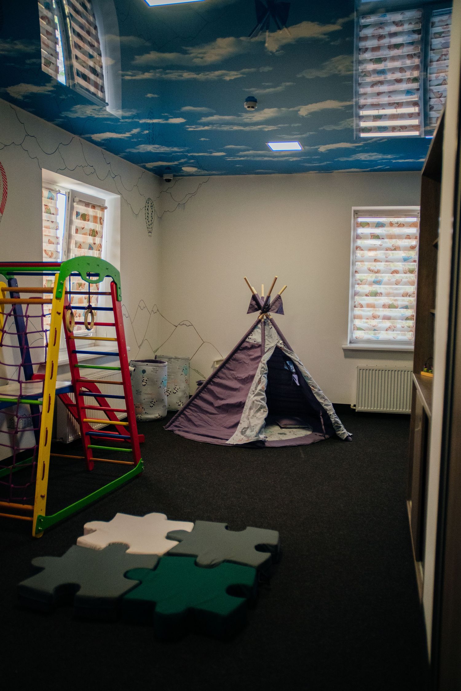 Colorful children's playroom with a teepee tent, jungle gym, puzzle floor mats, and sky-themed ceiling mural.