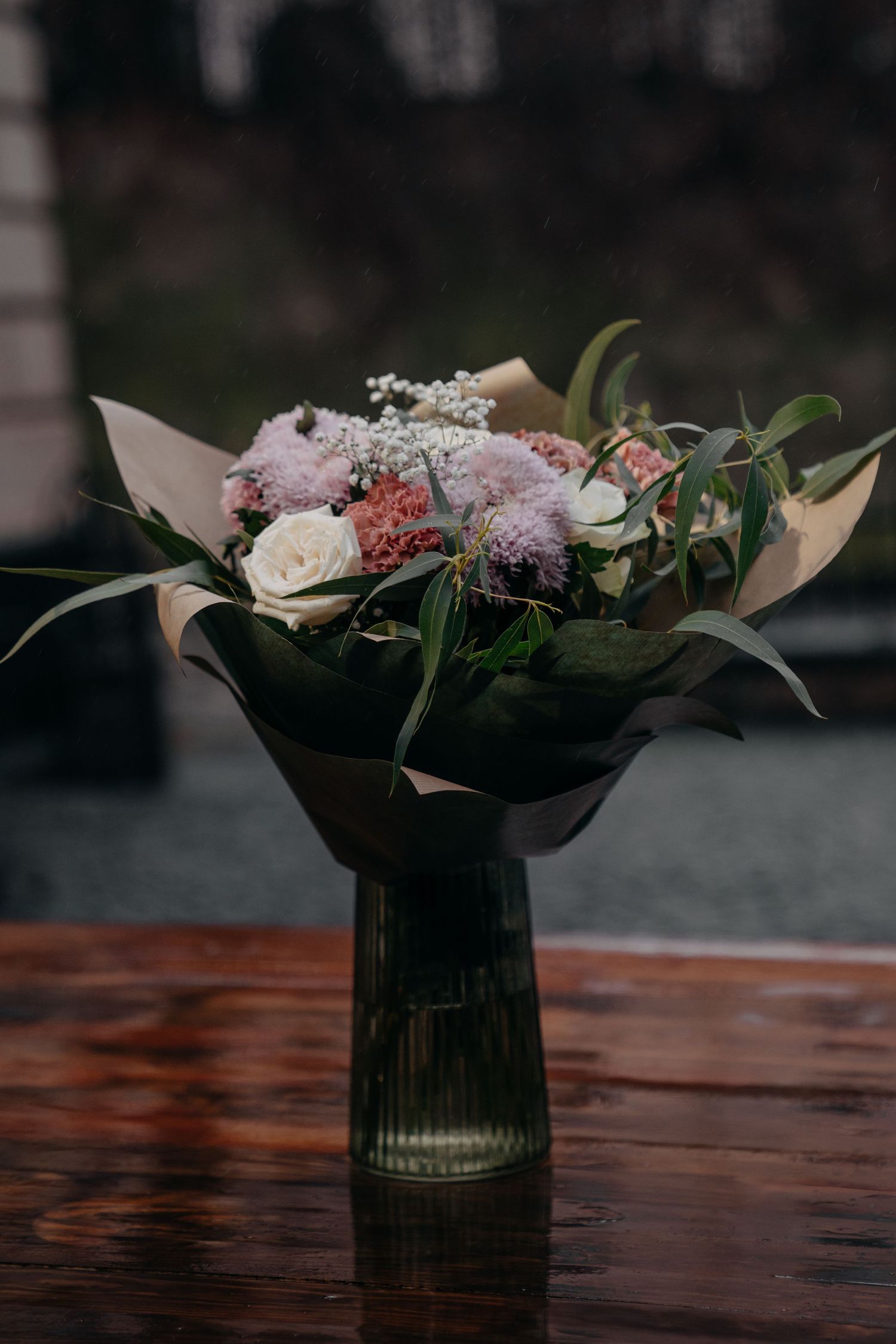 Elegant floral bouquet with pink chrysanthemums, white roses, and greenery in a dark glass vase on a wooden table.