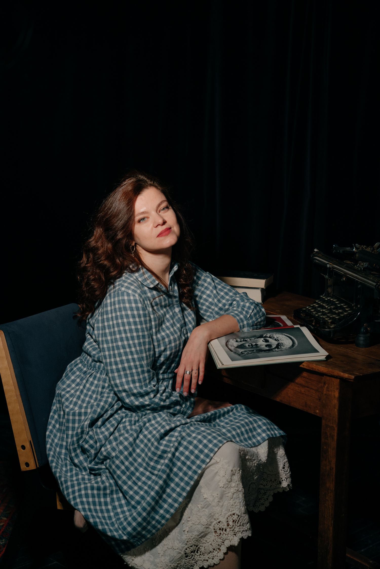 Young woman in green plaid dress sitting at vintage desk holding a book in dramatic dark studio lighting.
