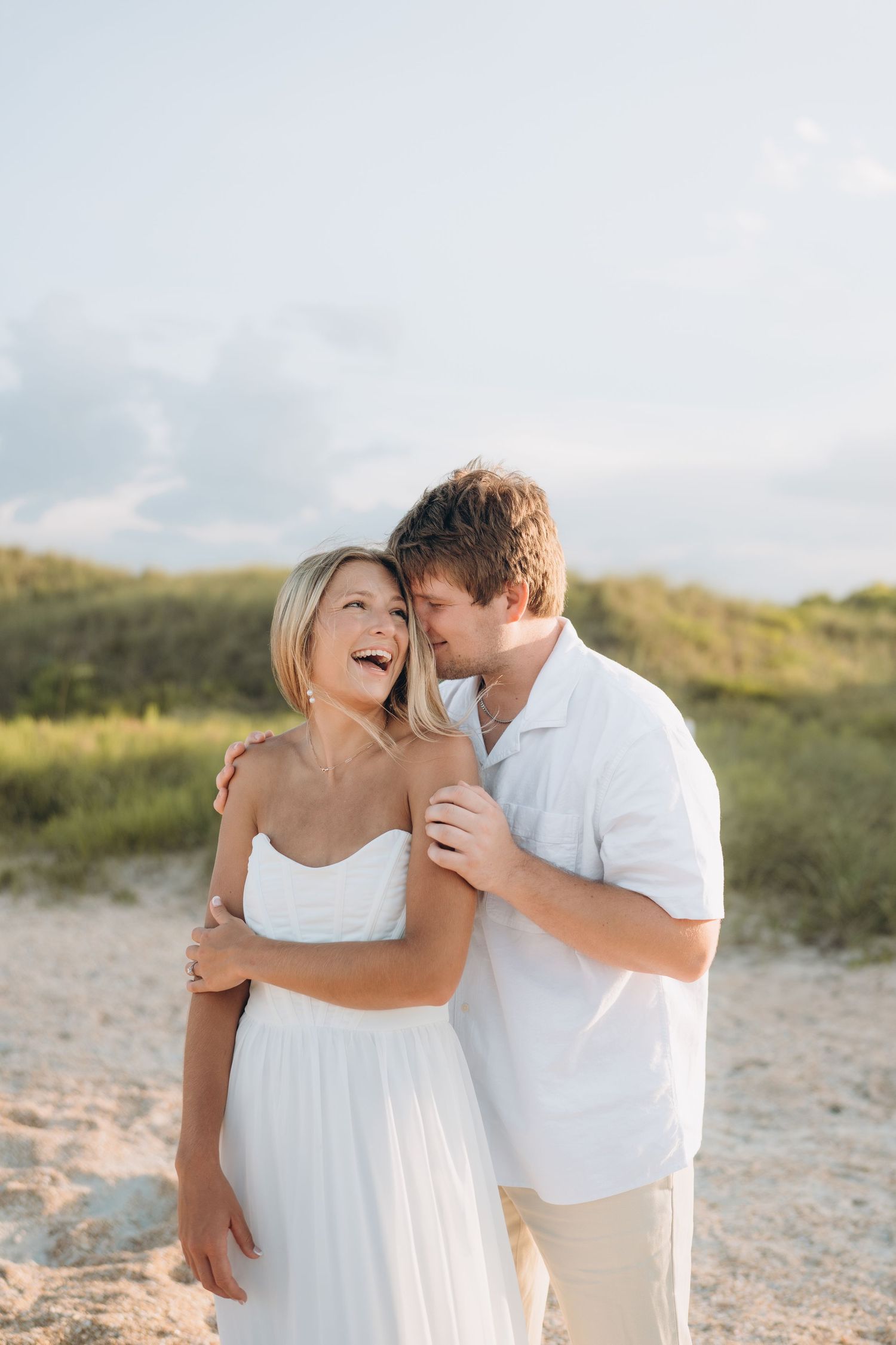 A couple shares an intimate moment on the beach wearing white outfits in soft sunlight.