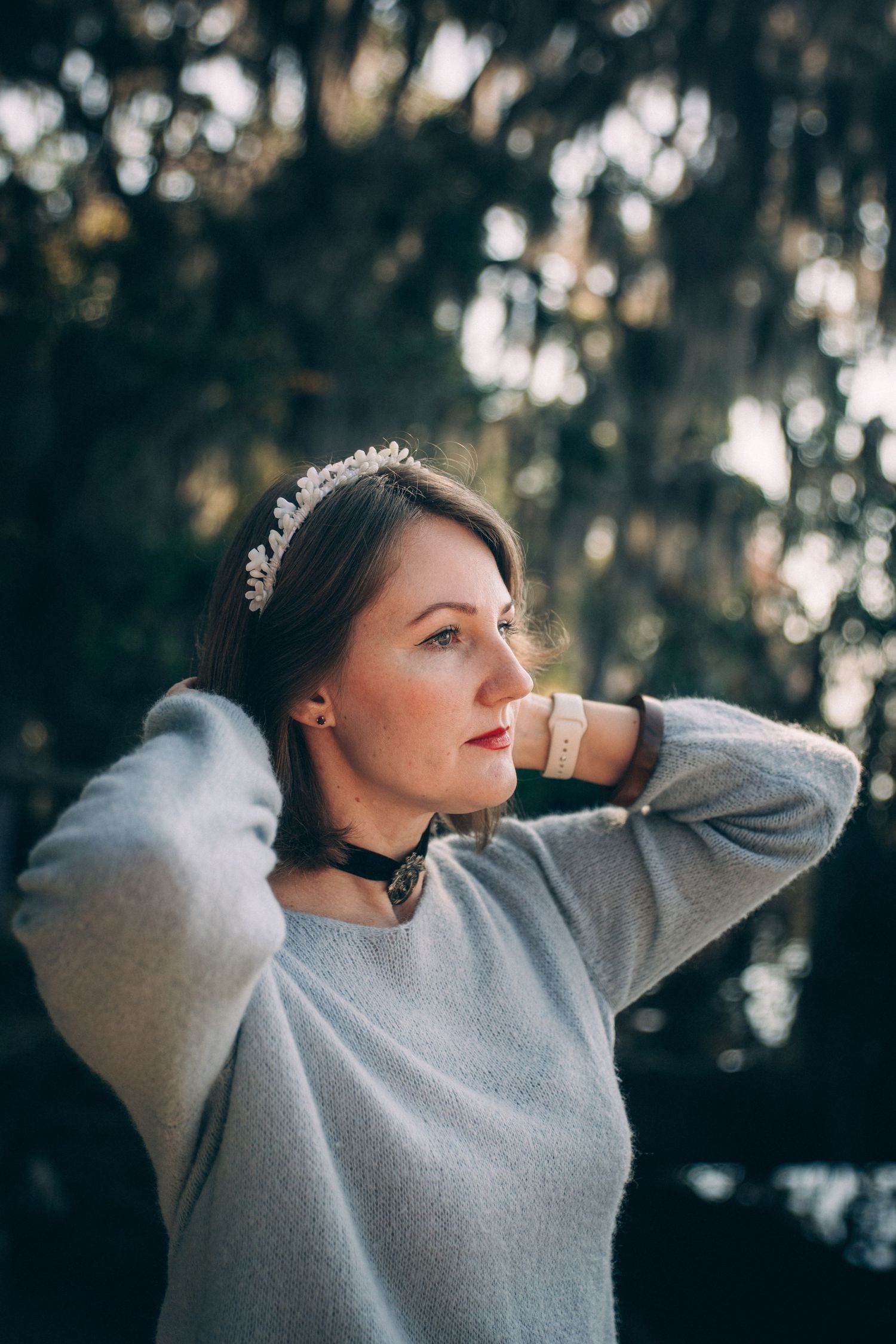 A figure in a light sweater and choker poses against a backdrop of moss-covered trees.