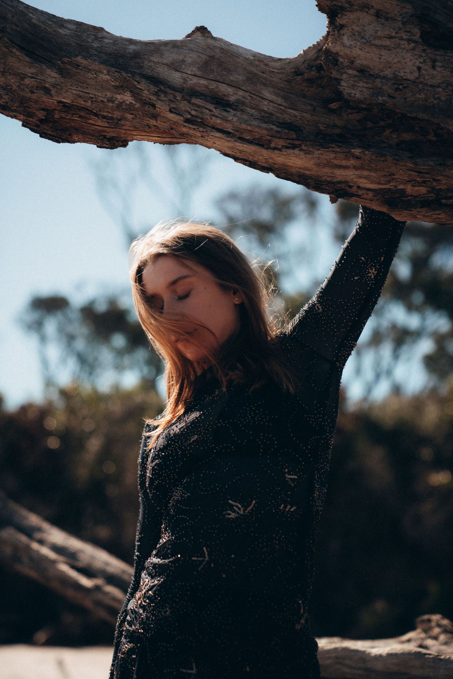 A figure in dark clothing leans against a weathered tree trunk in a moody outdoor setting with natural lighting.