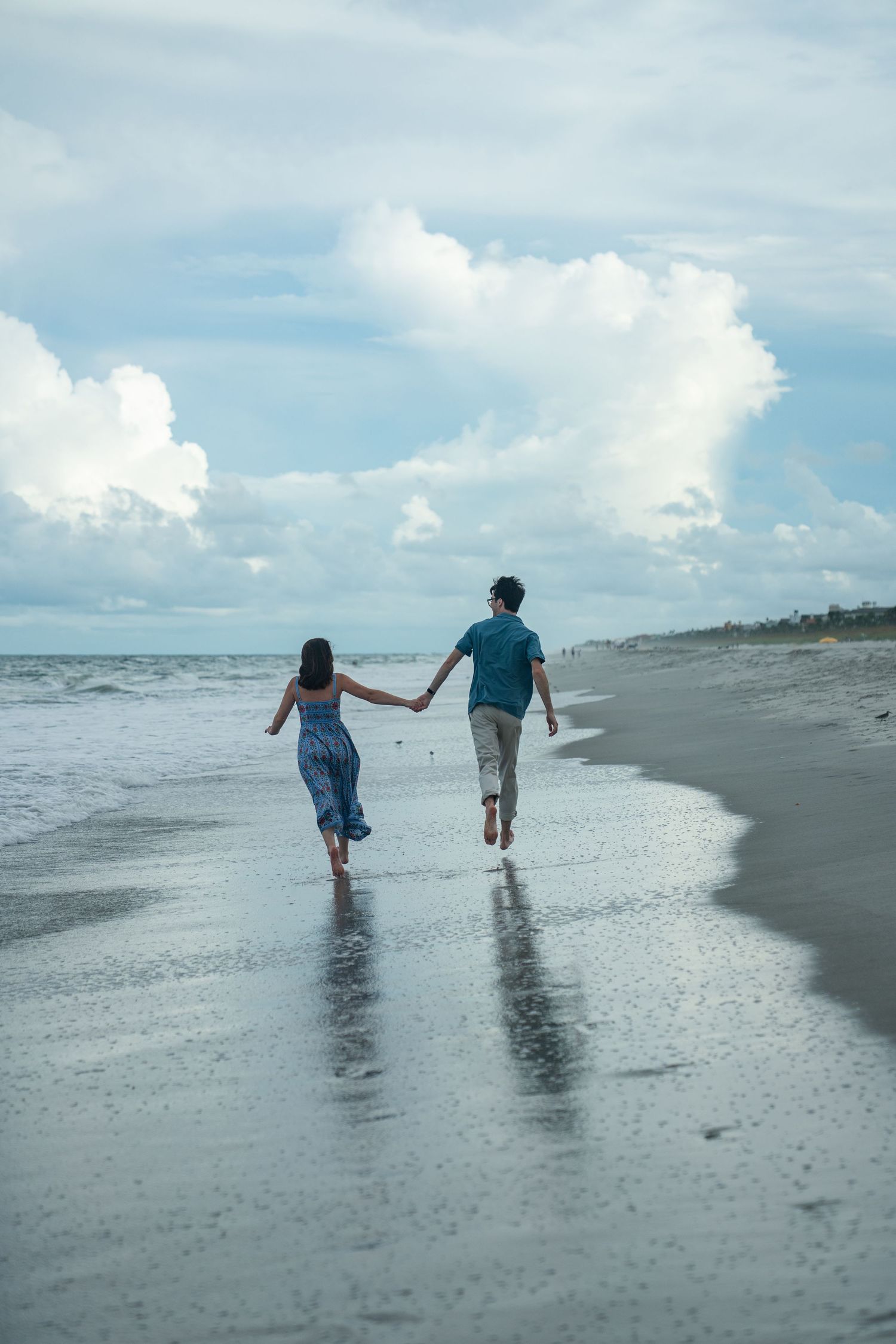A couple holding hands while running along the shoreline of a beach under a cloudy sky.