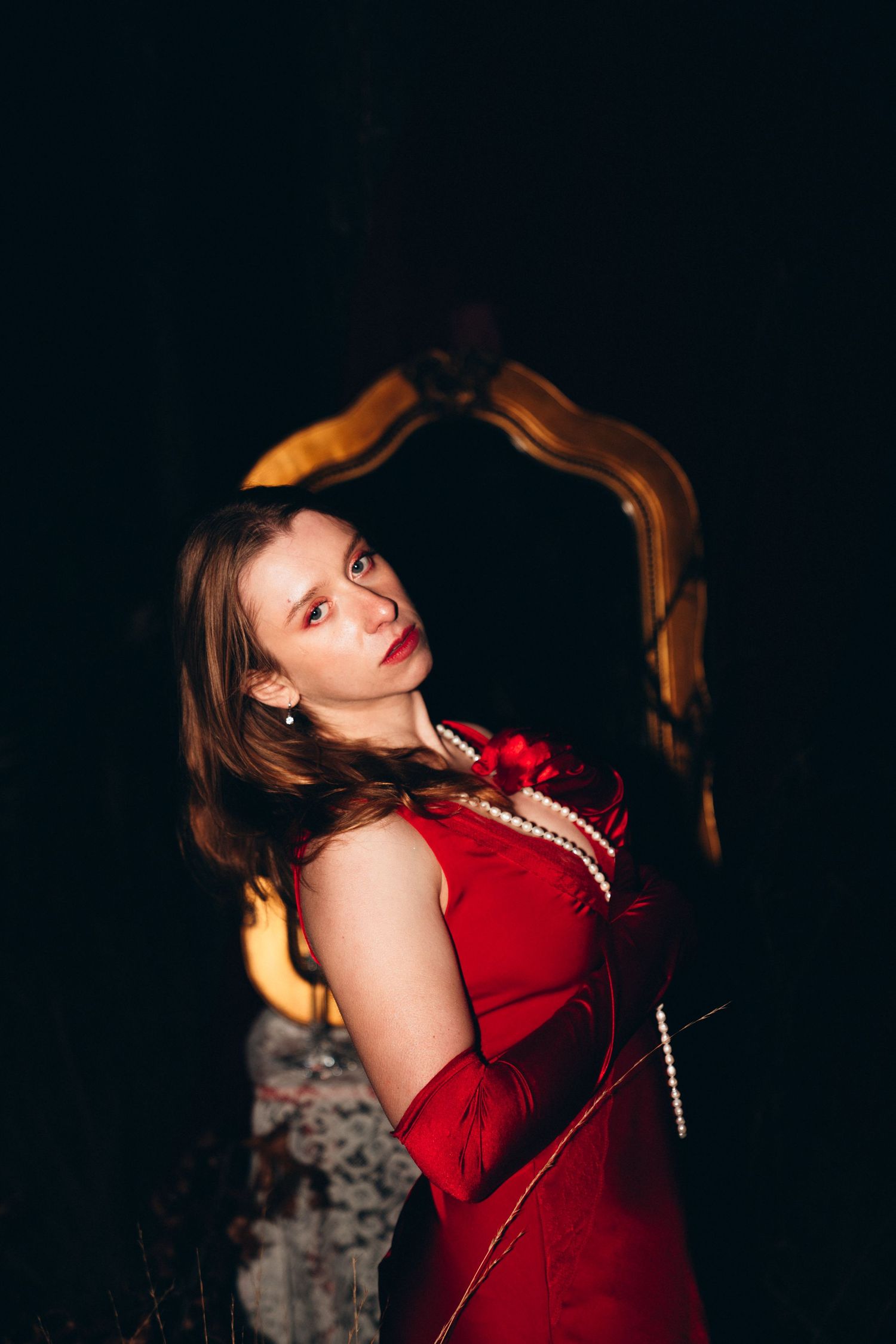 Woman in red dress and pearl necklace posing elegantly against dark background with ornate gold mirror frame.
