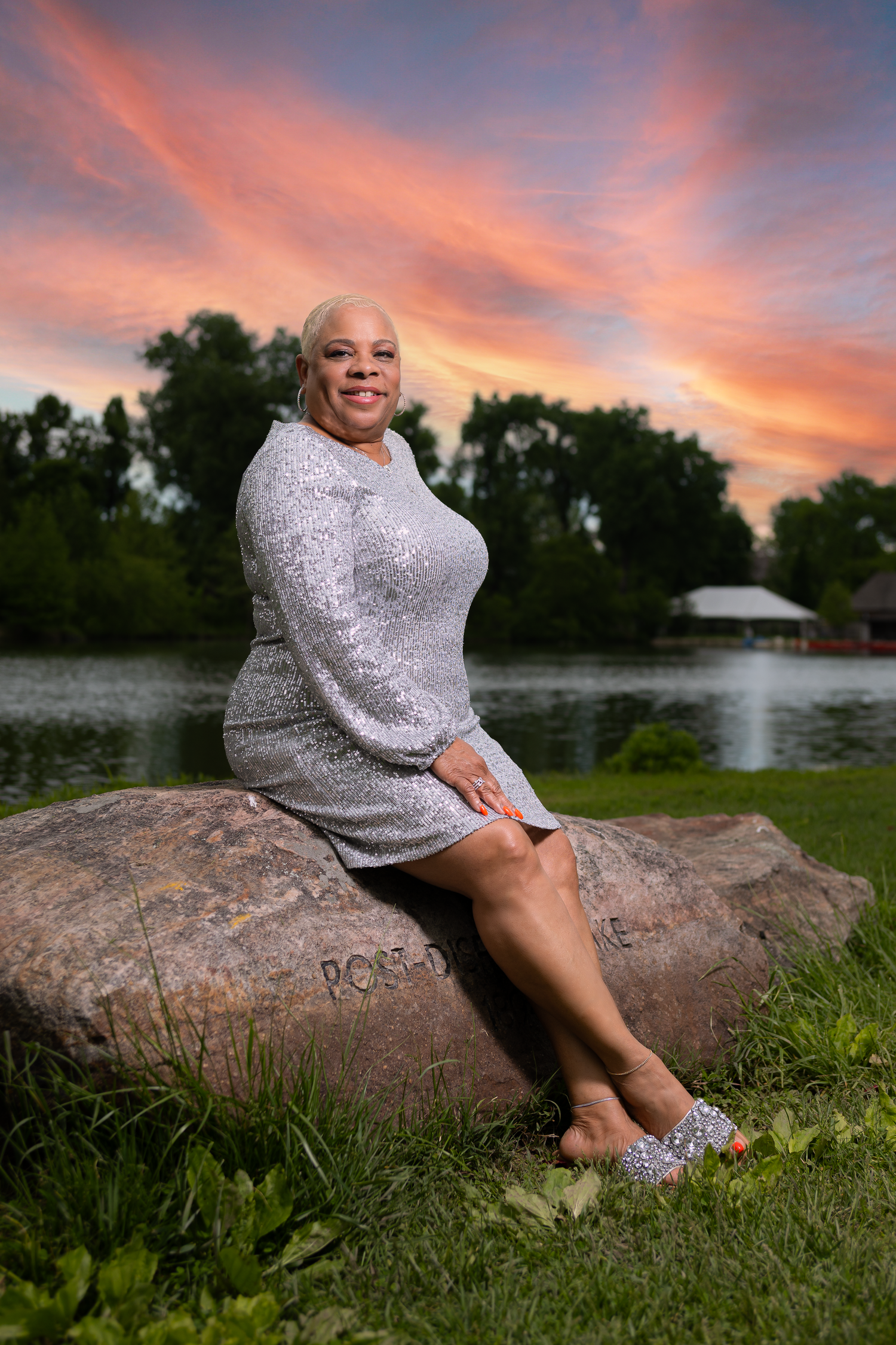 Person in sparkly dress sits on large rock by lake during vibrant sunset.