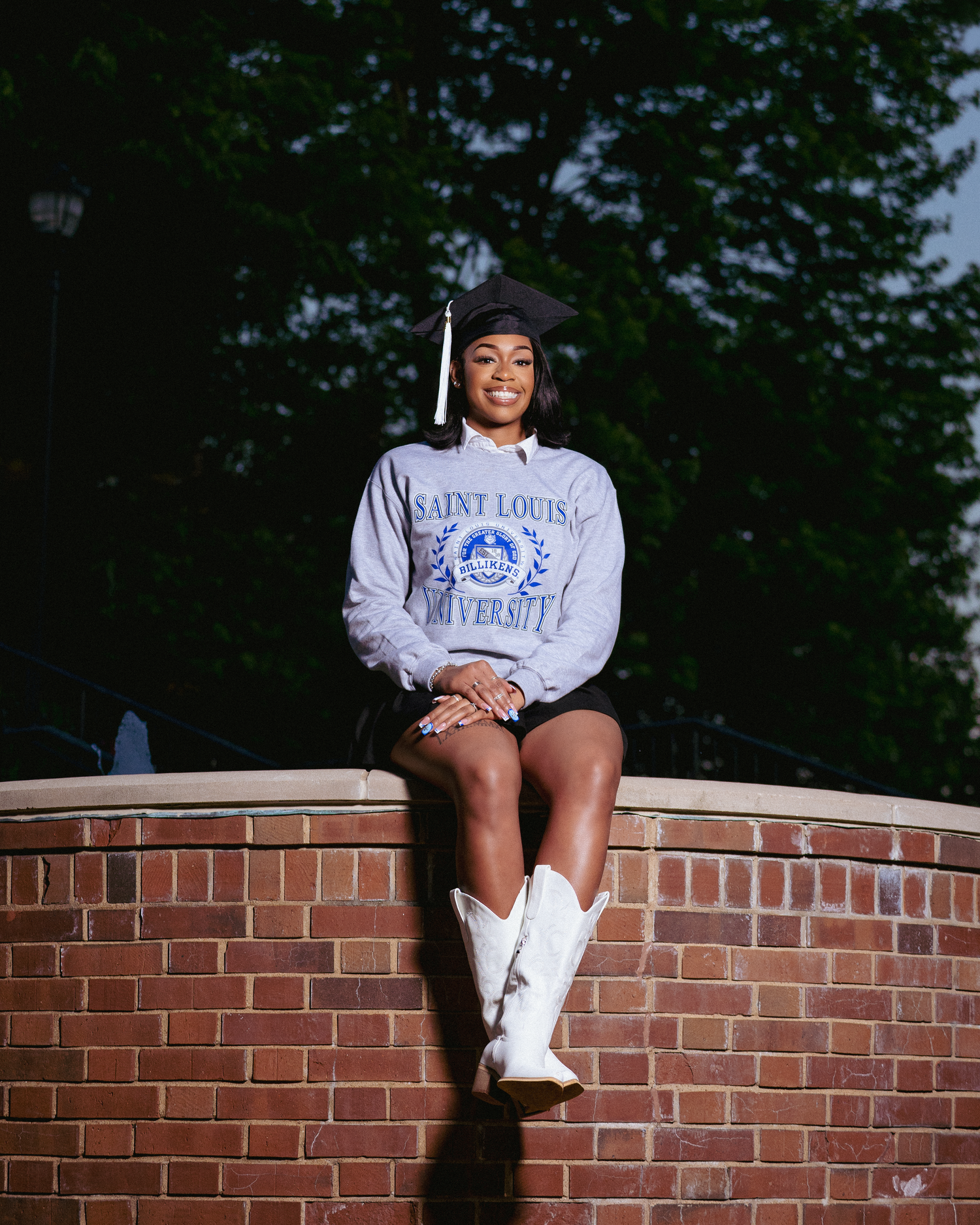 A young woman in a Saint Louis University sweatshirt and graduation cap sits on a brick wall wearing white cowboy boots.