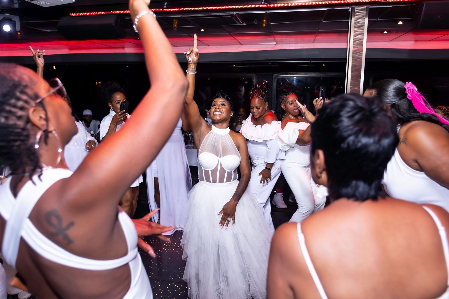 Celebratory scene of guests in white outfits dancing at a party under red lighting.