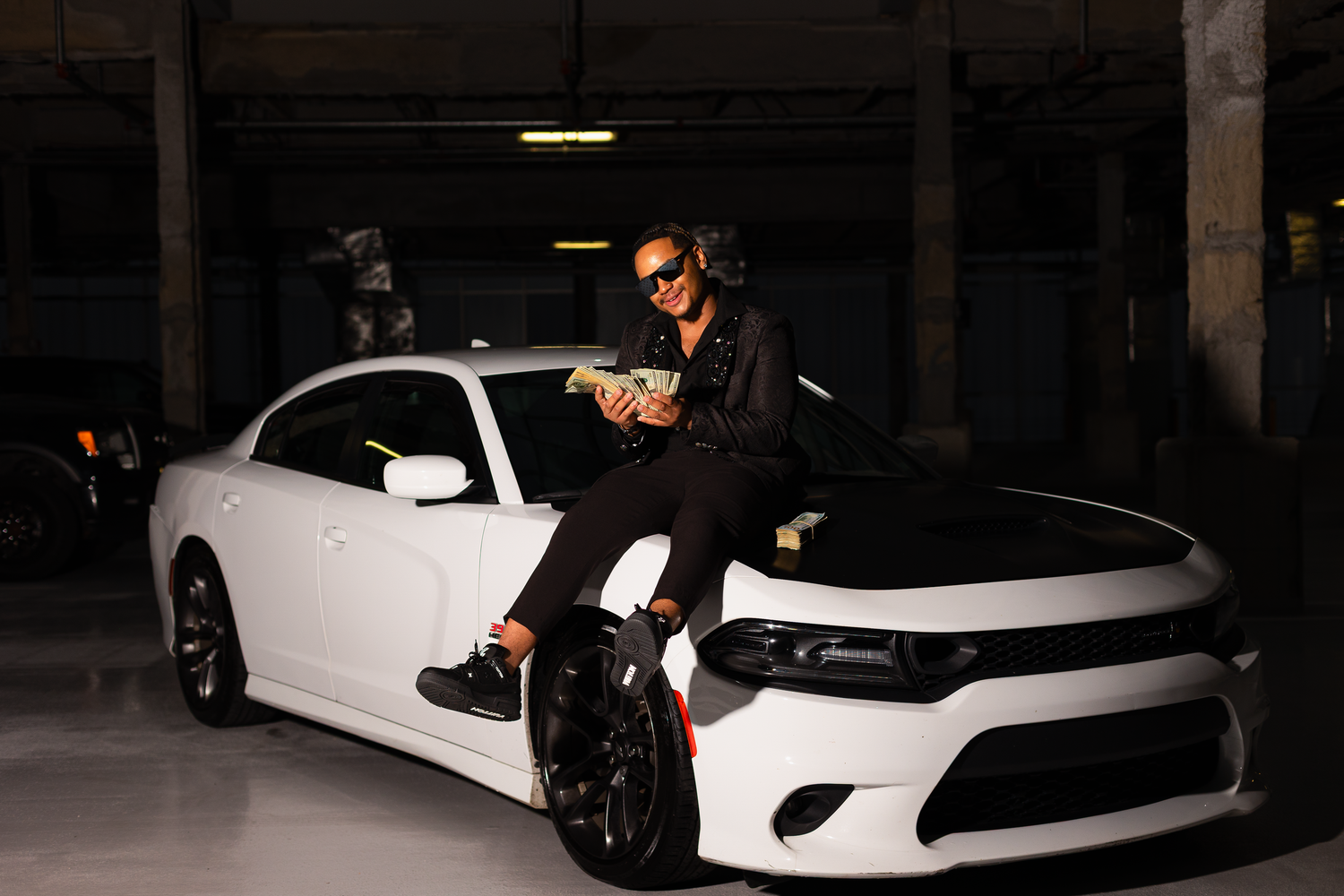 Person in black outfit sits on hood of white Dodge Charger in parking garage.