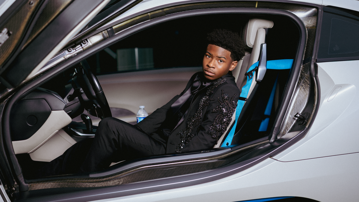 Young man in black jacket sitting in luxury sports car with blue accents and racing seat.