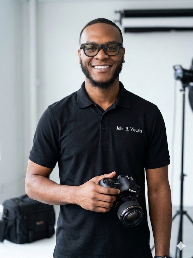 Smiling photographer holding a Canon camera wearing a black John B. Visuals polo shirt in a professional studio.